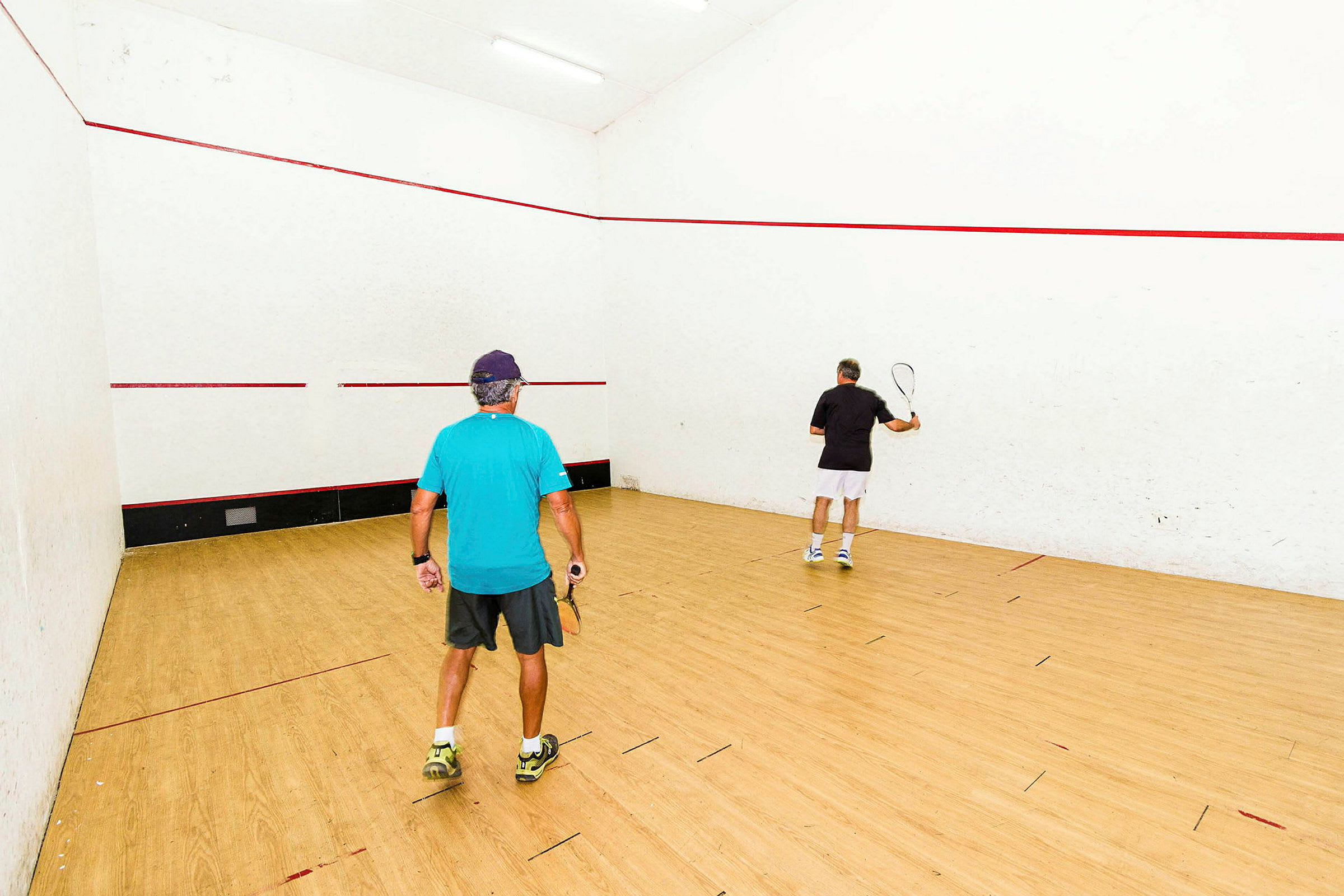 Två personer spelar squash på en inomhusbana med ljusa väggar och trägolv. (AI-genererad text)