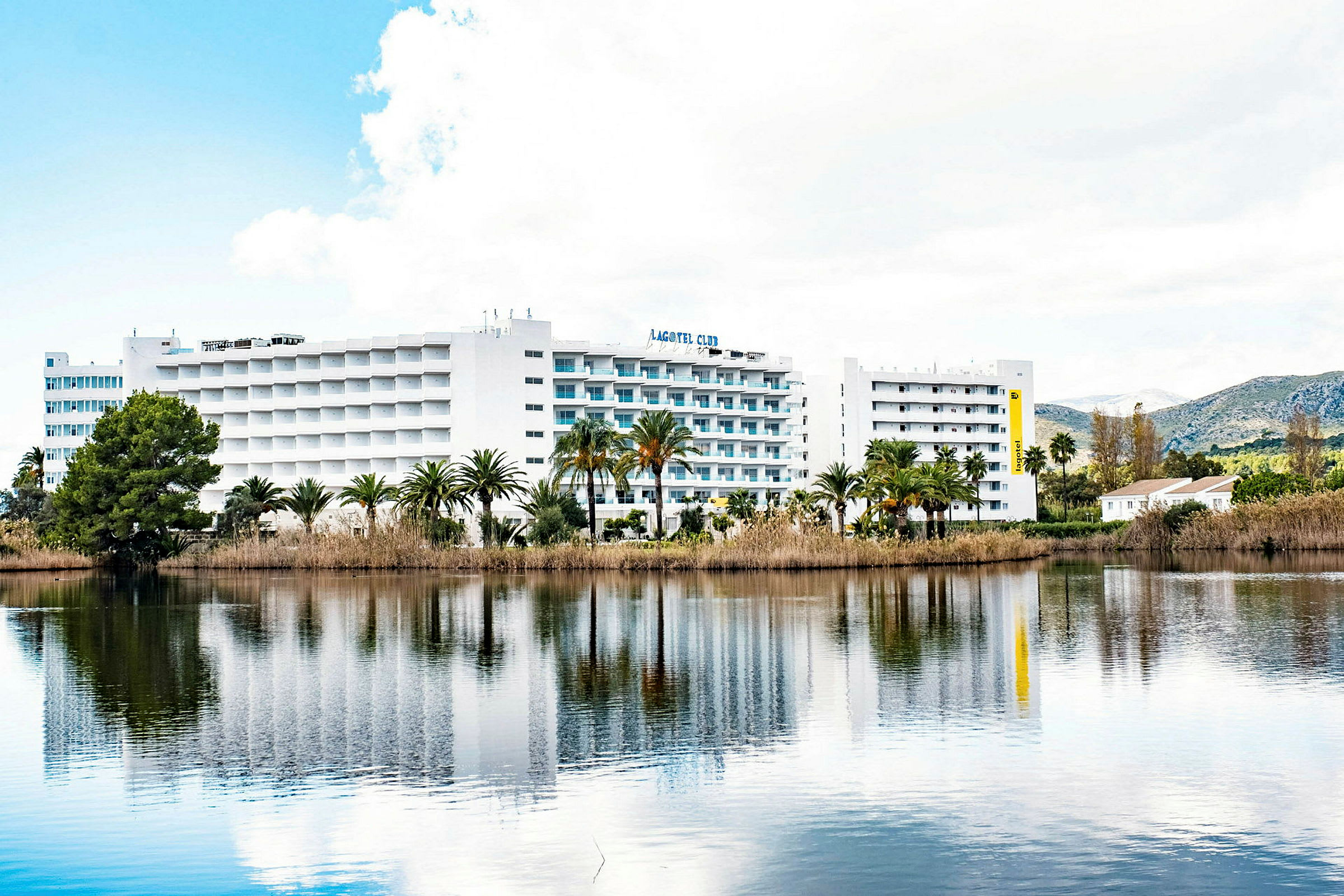 Ett hotellkomplex med vit fasad och balkonger reflekteras i en stilla sjö. Gröna palmer och vegetation omger byggnaden. (AI-genererad text)