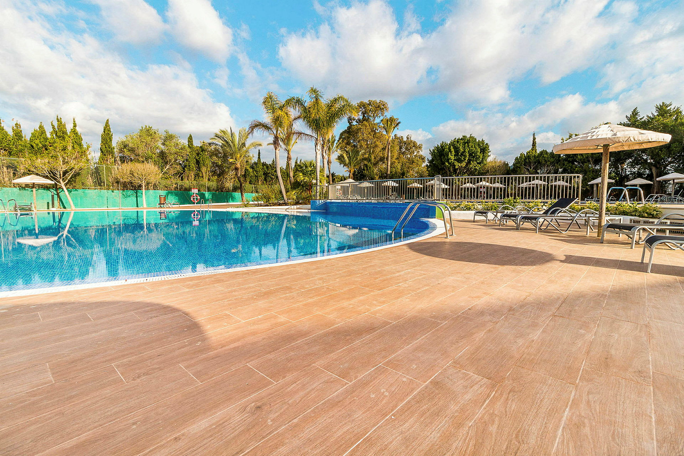 En utomhuspool med klarblått vatten, omgiven av solstolar och parasoller. Soligt väder med några moln. (AI-genererad text)