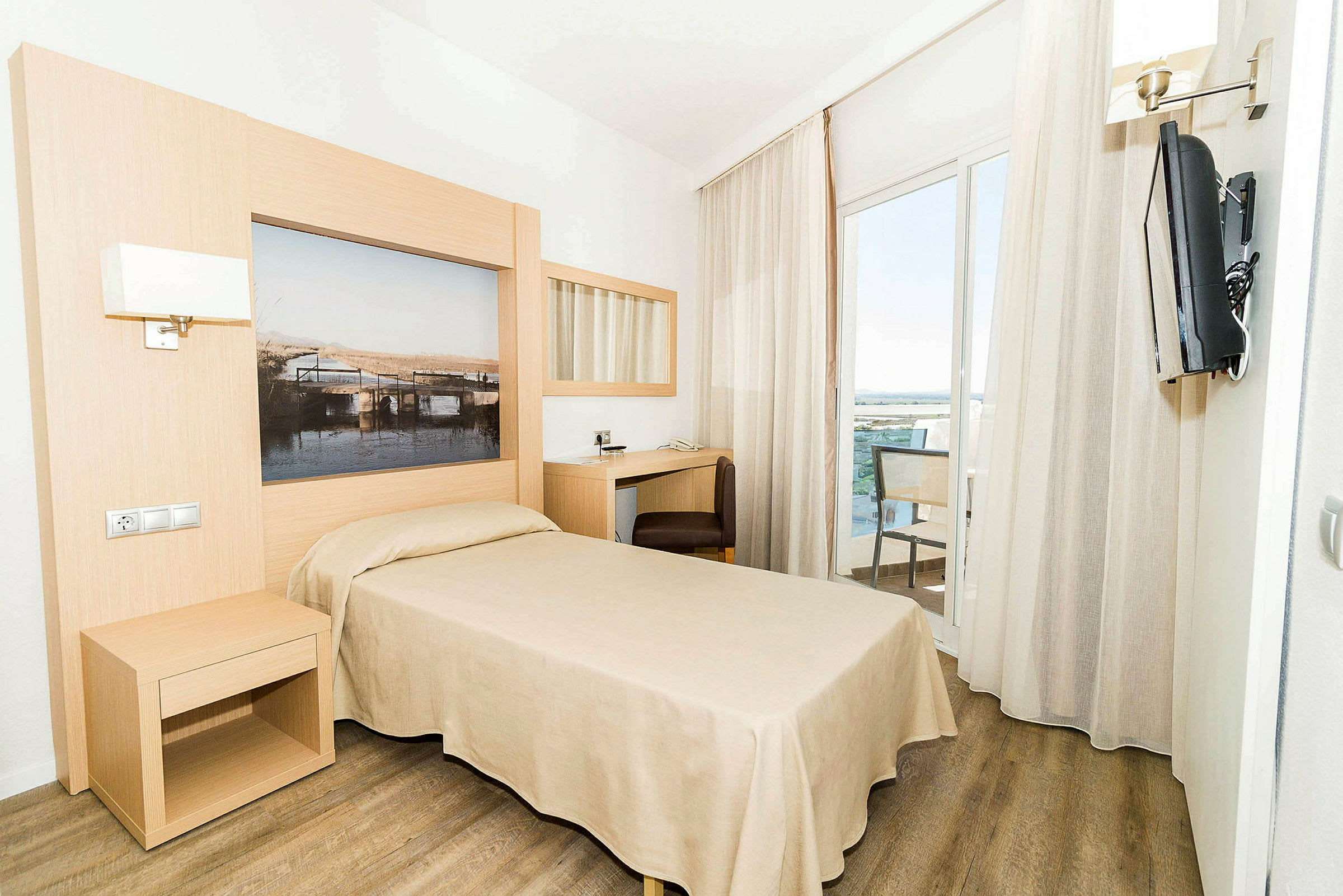 Ett hotellrum med en enkelsäng, ett skrivbord, en väggmonterad TV och ett stort fönster som leder ut till en balkong. (AI-genererad text)