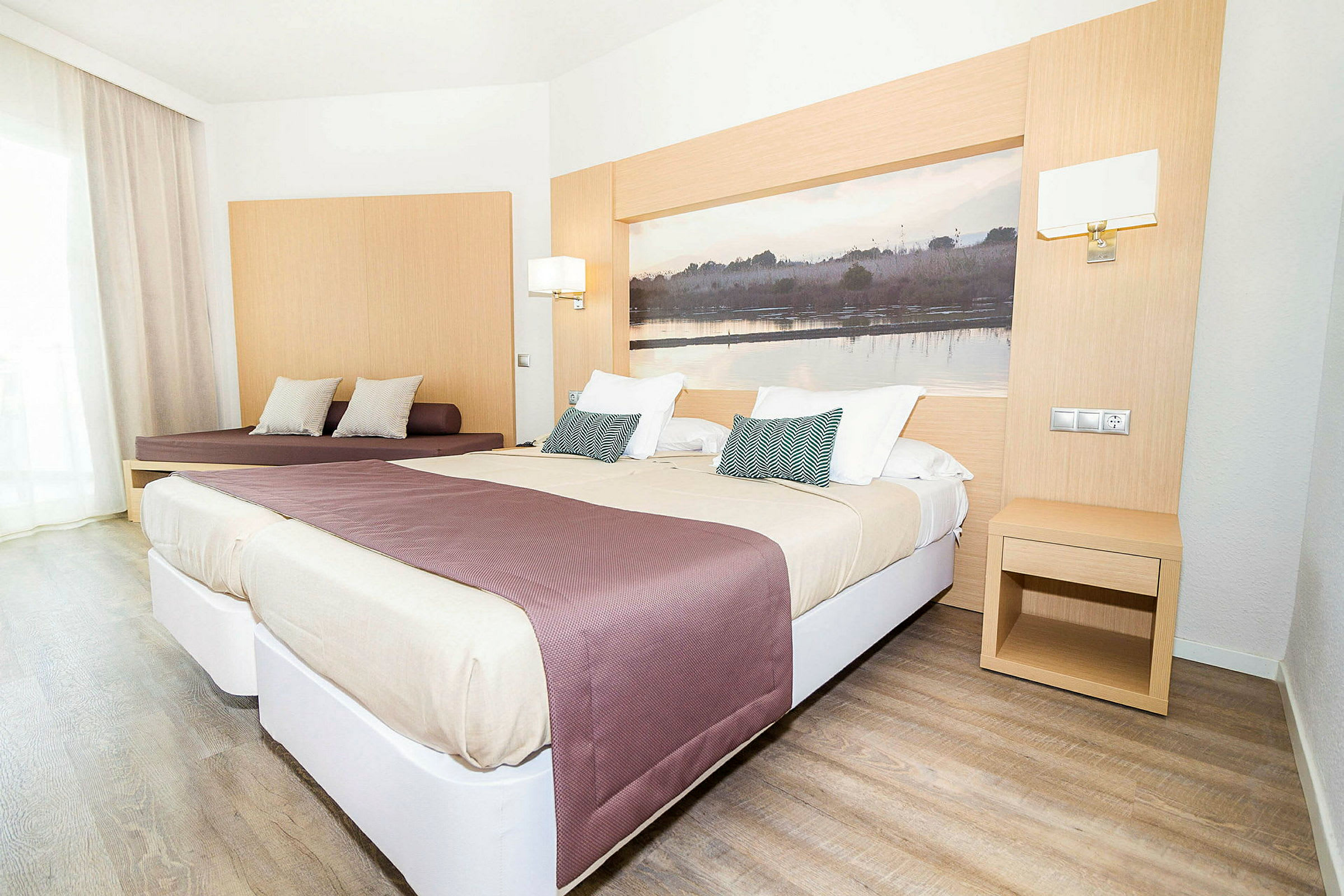 Ett ljust hotellrum med två sängar och en sittplats. Sängarna är bäddade med beige och lila överkast, och en stor bild pryder väggen bakom sängarna. (AI-genererad text)
