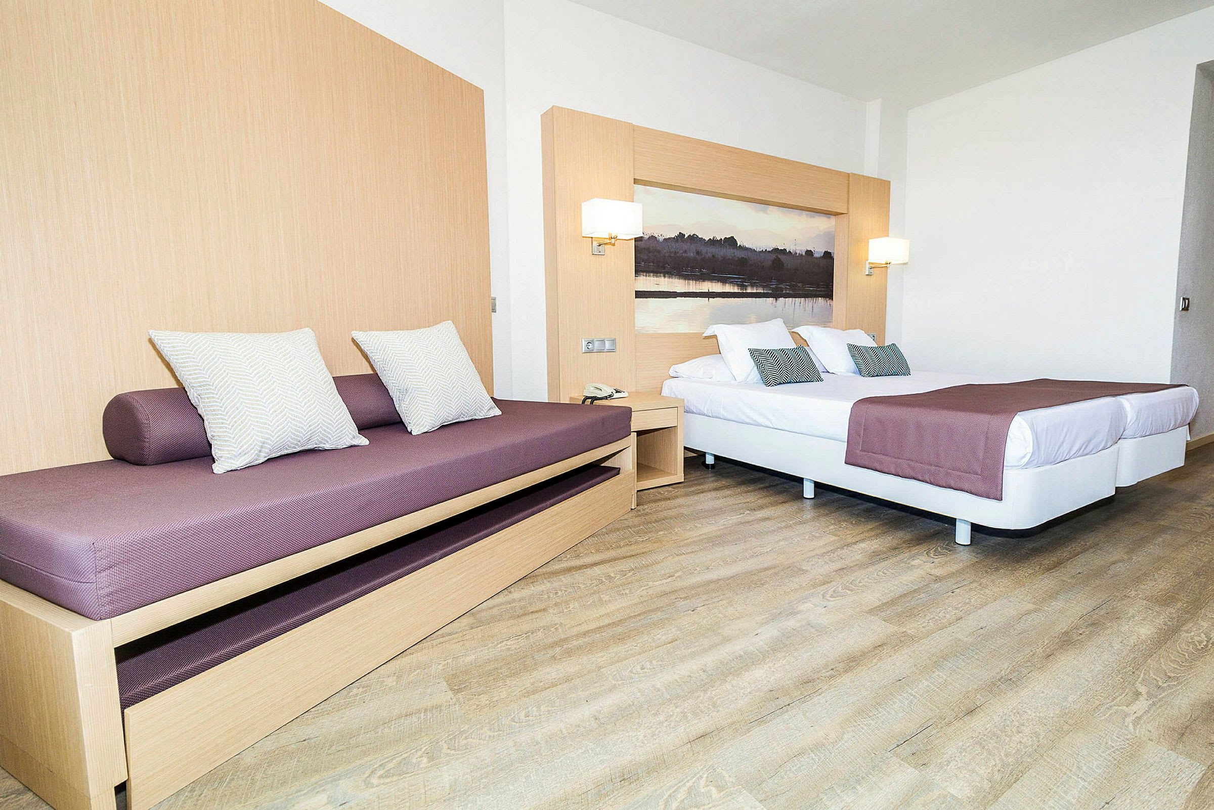 En hotellrumsinteriör med en bäddsoffa och en dubbelsäng med lila överkast och prydnadskuddar. (AI-genererad text)