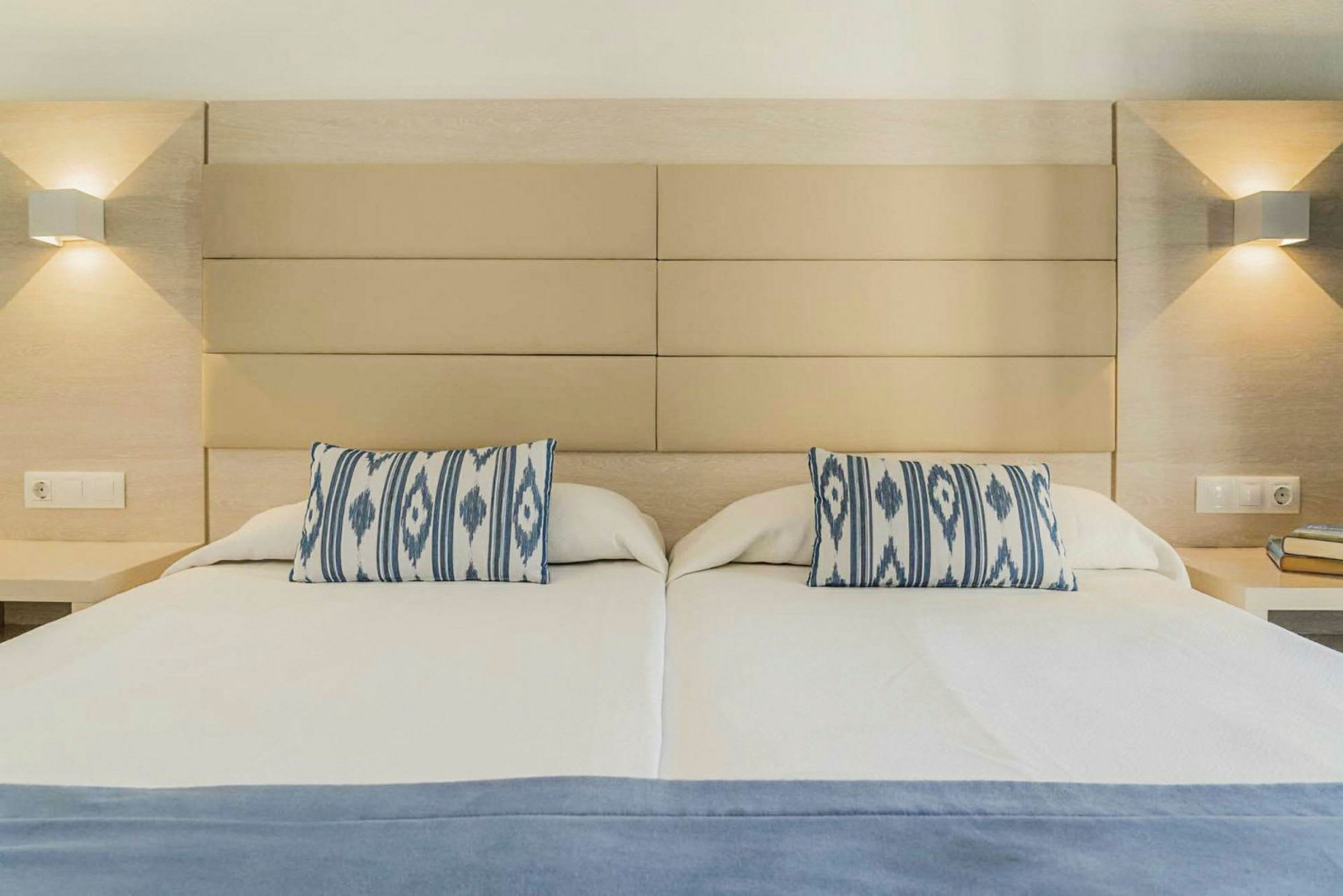 Ett hotellrum med en dubbelsäng. Sängen har två dekorativa kuddar med blå och vita mönster och en blå filt över sängen. Ljusa vägglampor finns på båda sidor av sängen. (AI-genererad text)