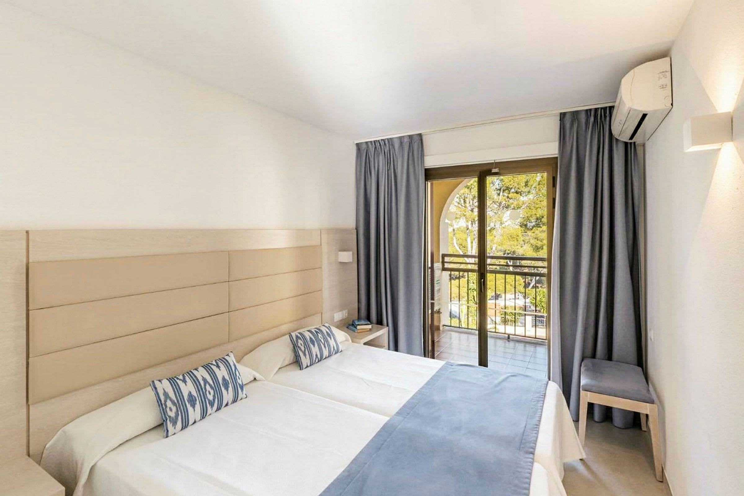 Ett hotellrum med två enkelsängar, ljus inredning, blåa sängöverdrag och kuddar, samt en balkong med öppna glasdörrar. (AI-genererad text)