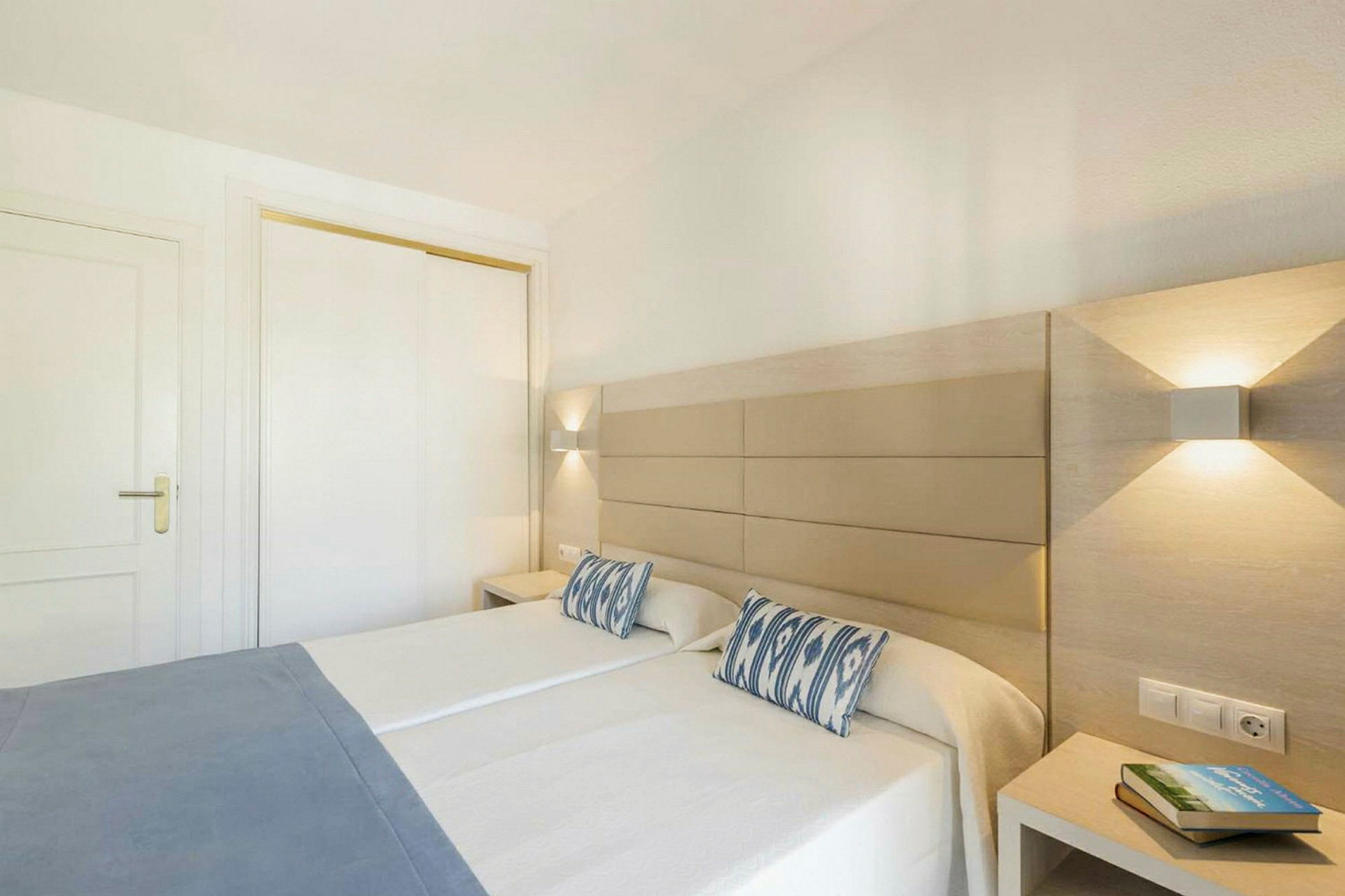 Två enkelsängar med vita sängkläder och blå dekorkuddar i ett modernt hotellrum. Ljus inredning med vägglampor vid sänggavlarna och ett nattduksbord med en bok. (AI-genererad text)