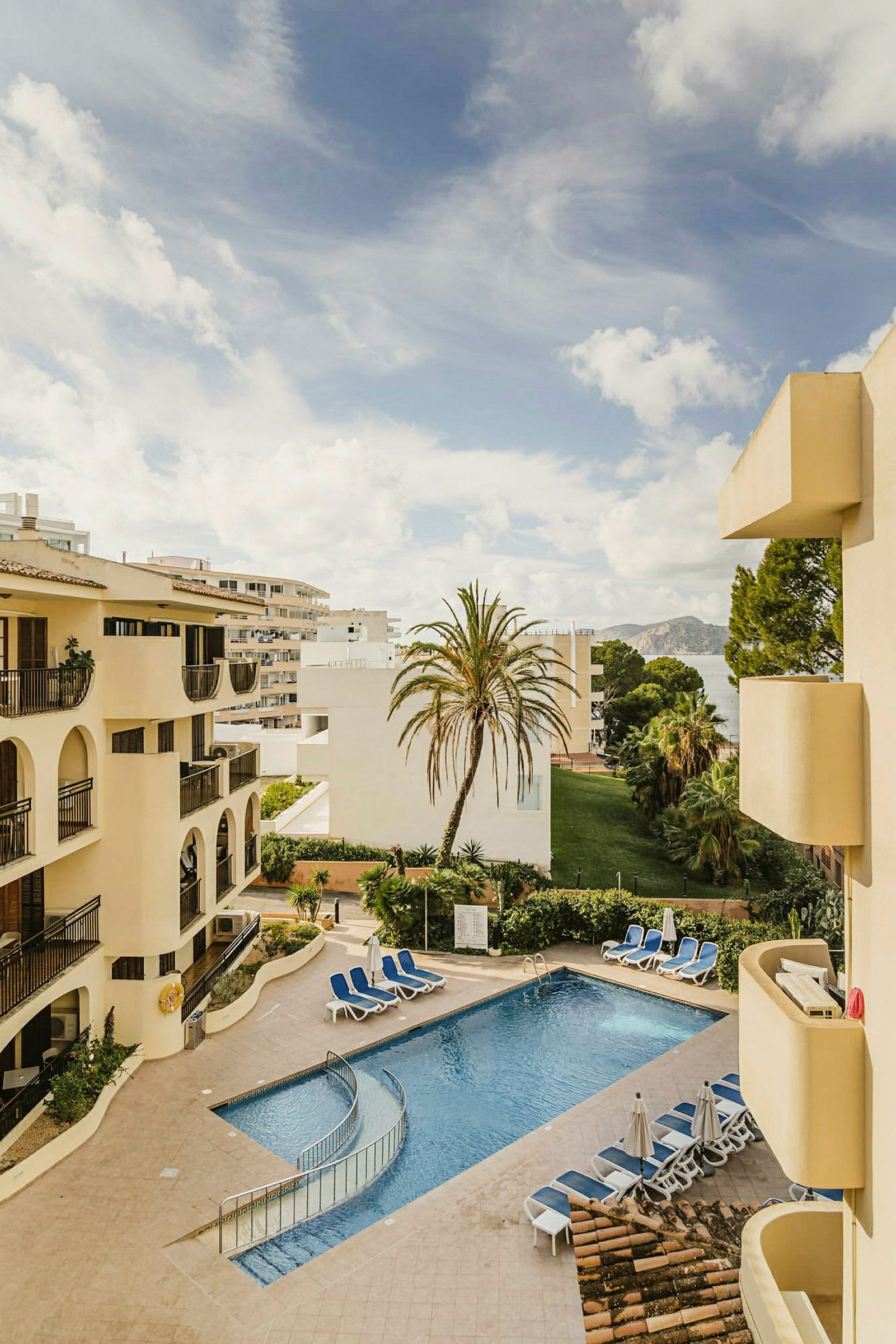 En utsikt över ett hotellområdes pool med omgivande byggnader, gröna områden, solstolar och en palm. (AI-genererad text)