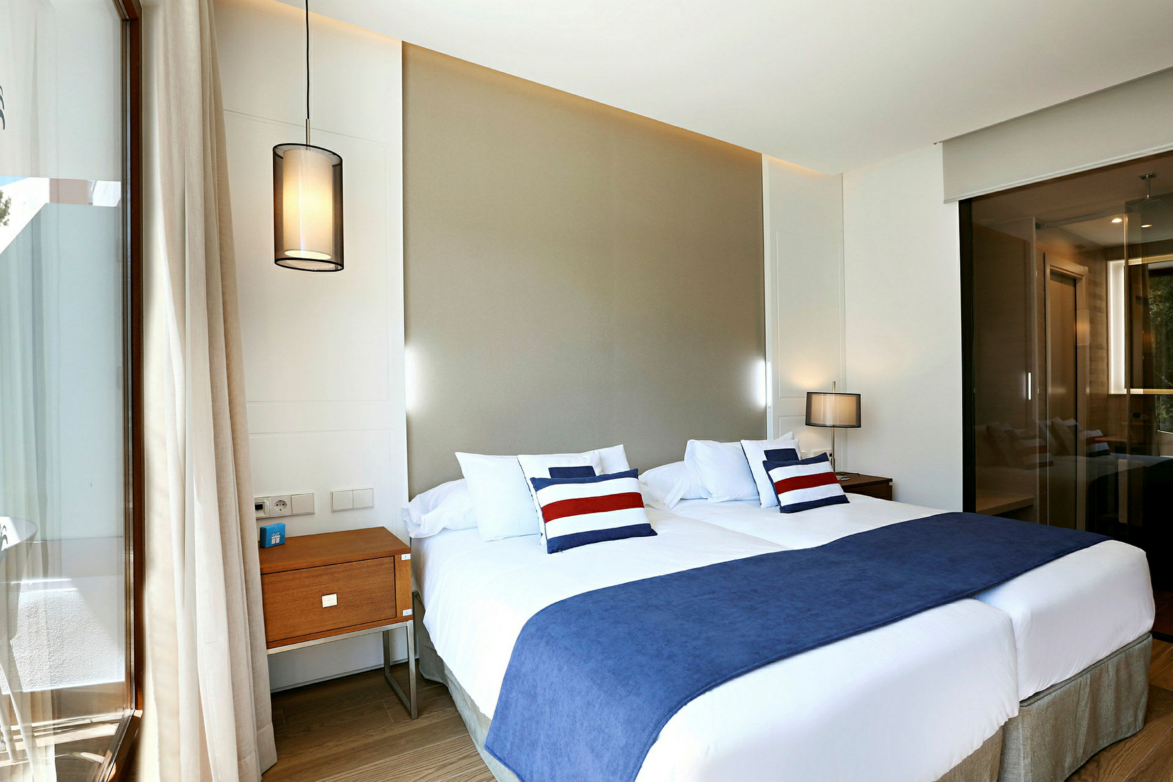 Ett hotellrum med två enkelsängar, blå överkast och dekorativa kuddar. Inredningen inkluderar hängande lampor och ett nattduksbord. (AI-genererad text)