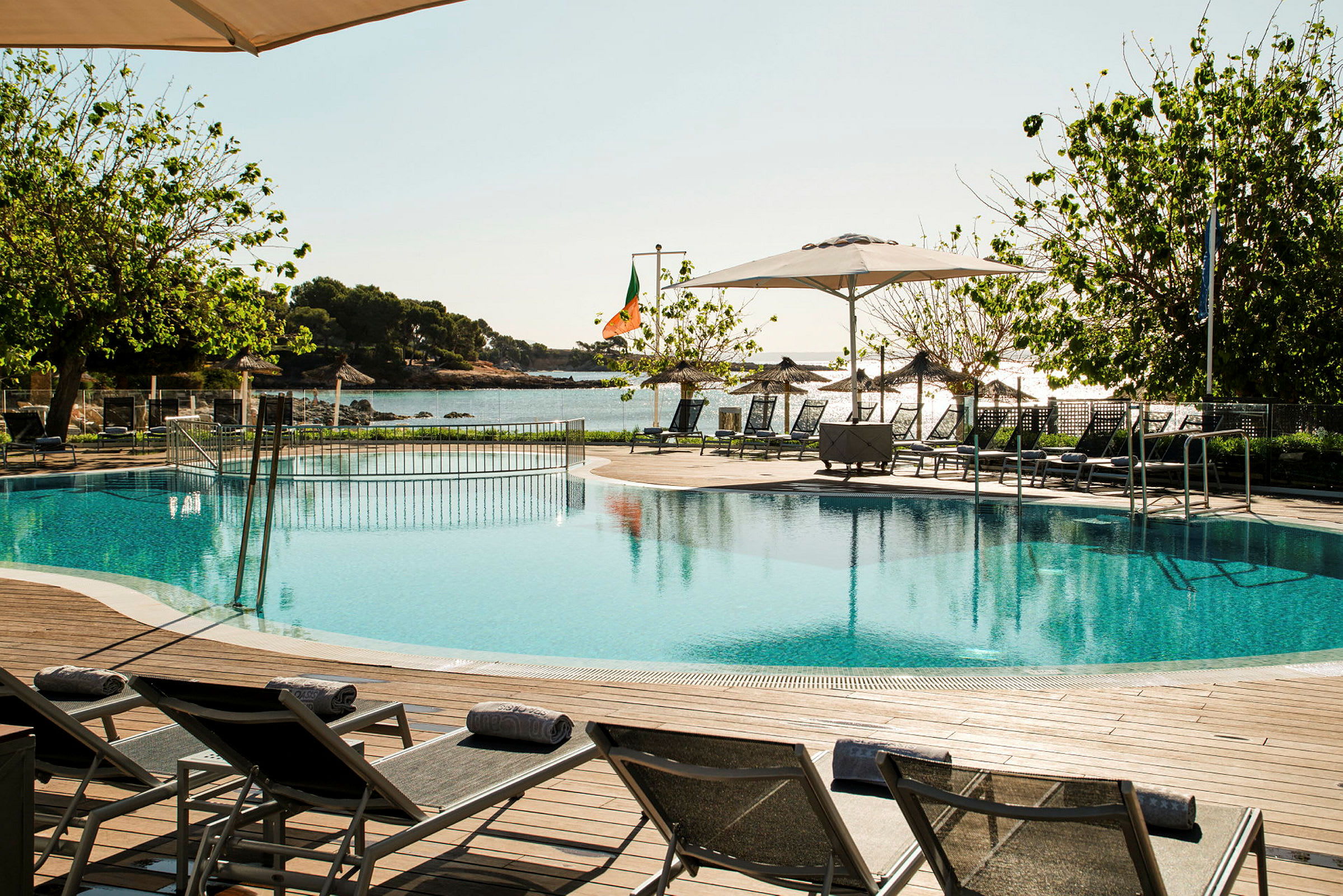 Pool med solsängar, träd och hav i bakgrunden. (AI-genererad text)