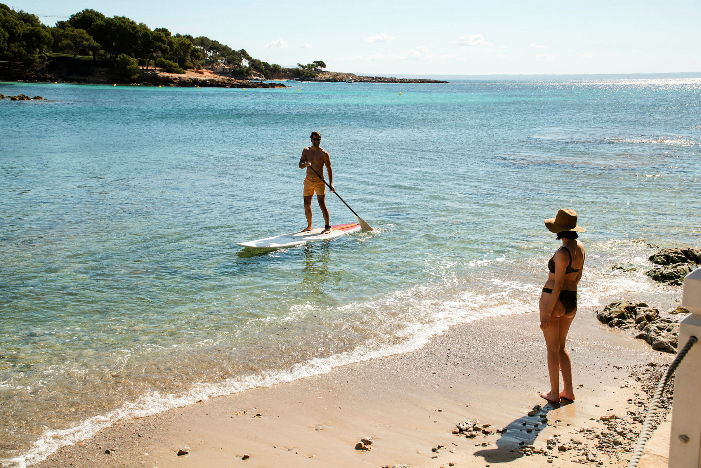 En person står på en paddleboard i havet nära stranden, medan en annan person står på sandstranden med utsikt mot vattnet. (AI-genererad text)