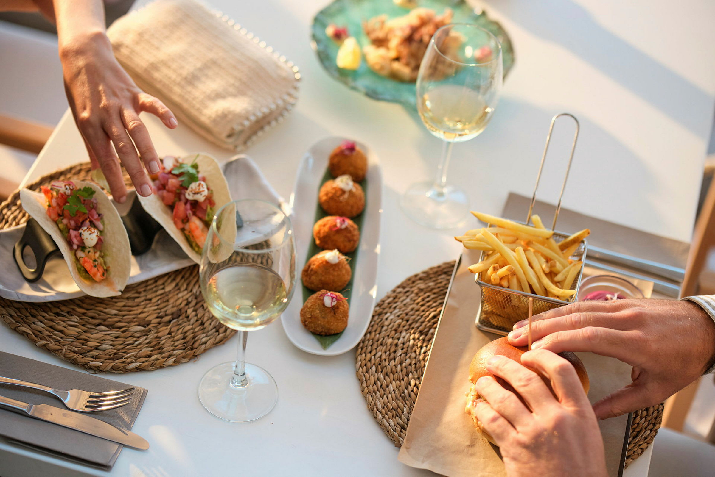 Närbild på en bordssituation med mat och dryck, inklusive tacos, croquetas, pommes frites, en hamburgare och vinglas. (AI-genererad text)