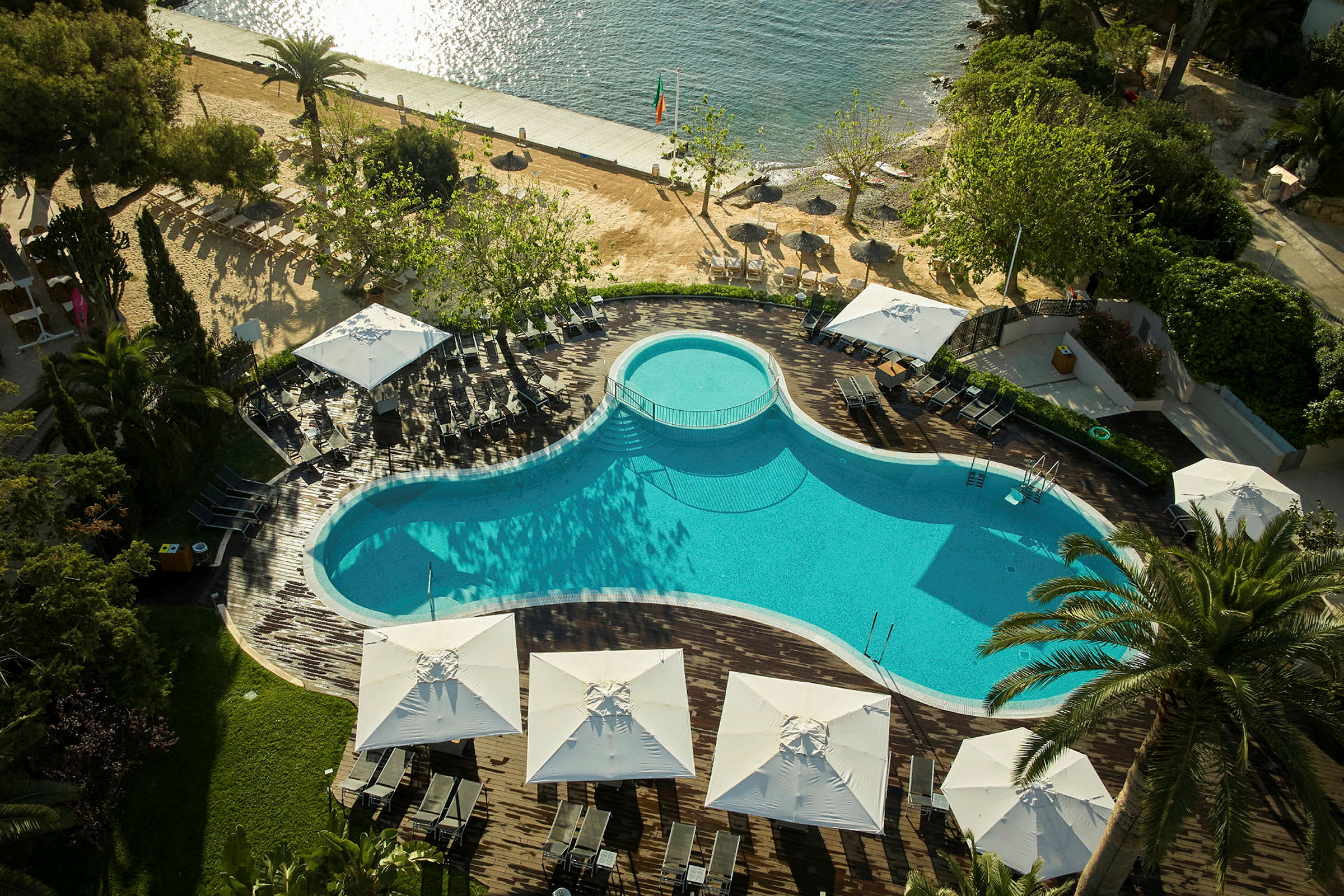 En pool omgiven av solstolar och parasoller med sandstrand och hav i bakgrunden. (AI-genererad text)