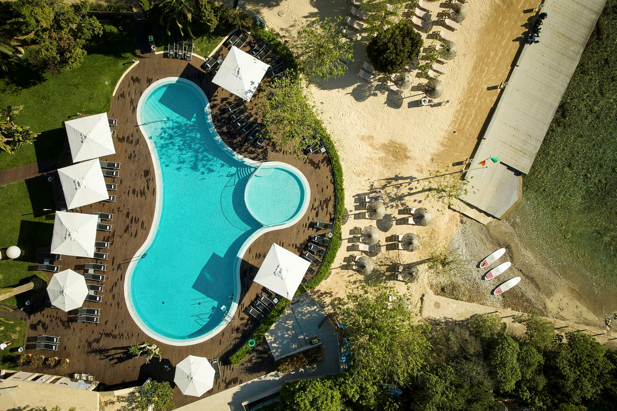 Luftbild över en pool omgiven av solstolar och parasoller, med en angränsande strand och brygga. (AI-genererad text)