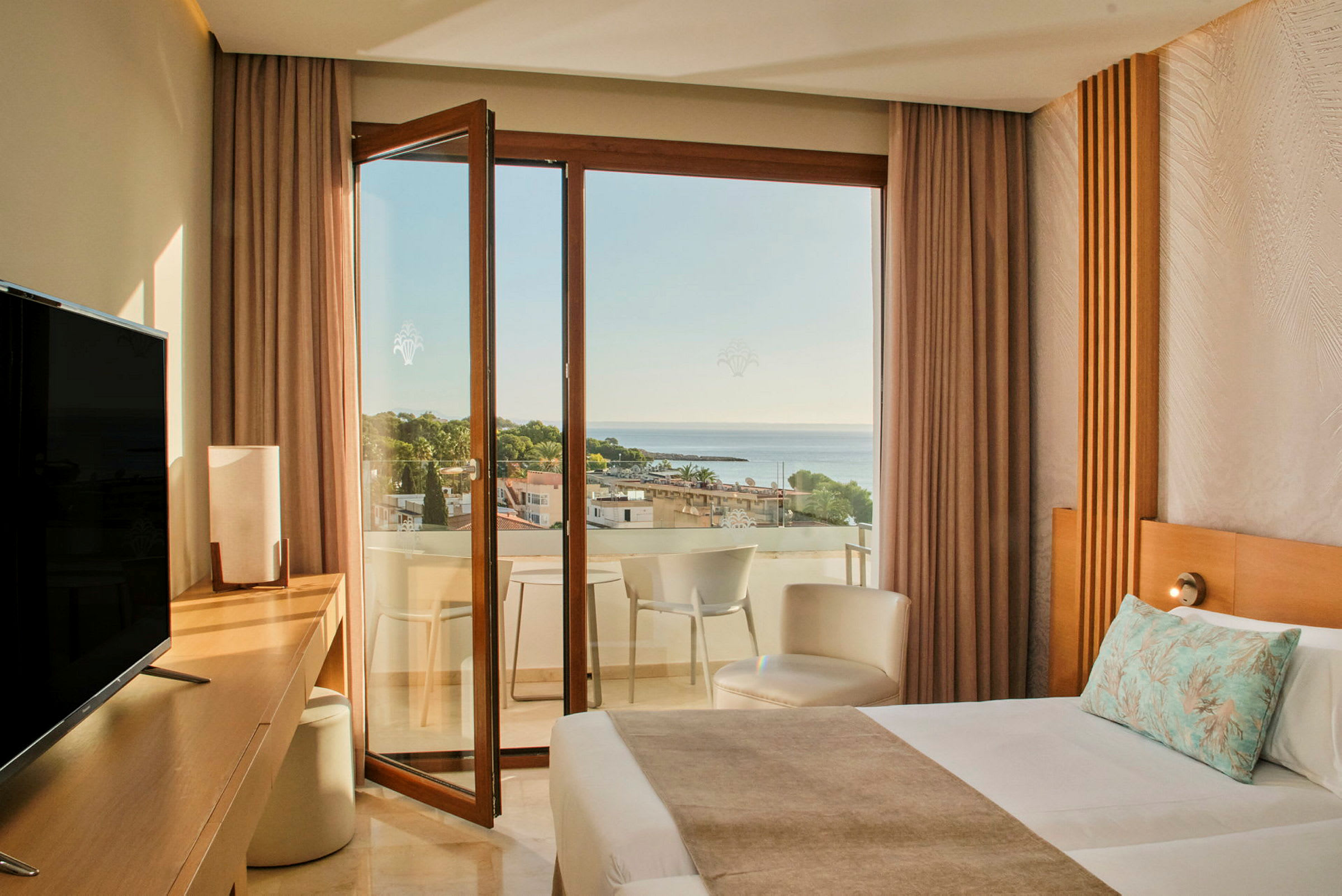 Ett hotellrum med en säng, skrivbord, TV och en öppen balkongdörr som leder till en balkong med utsikt över havet och träd. (AI-genererad text)