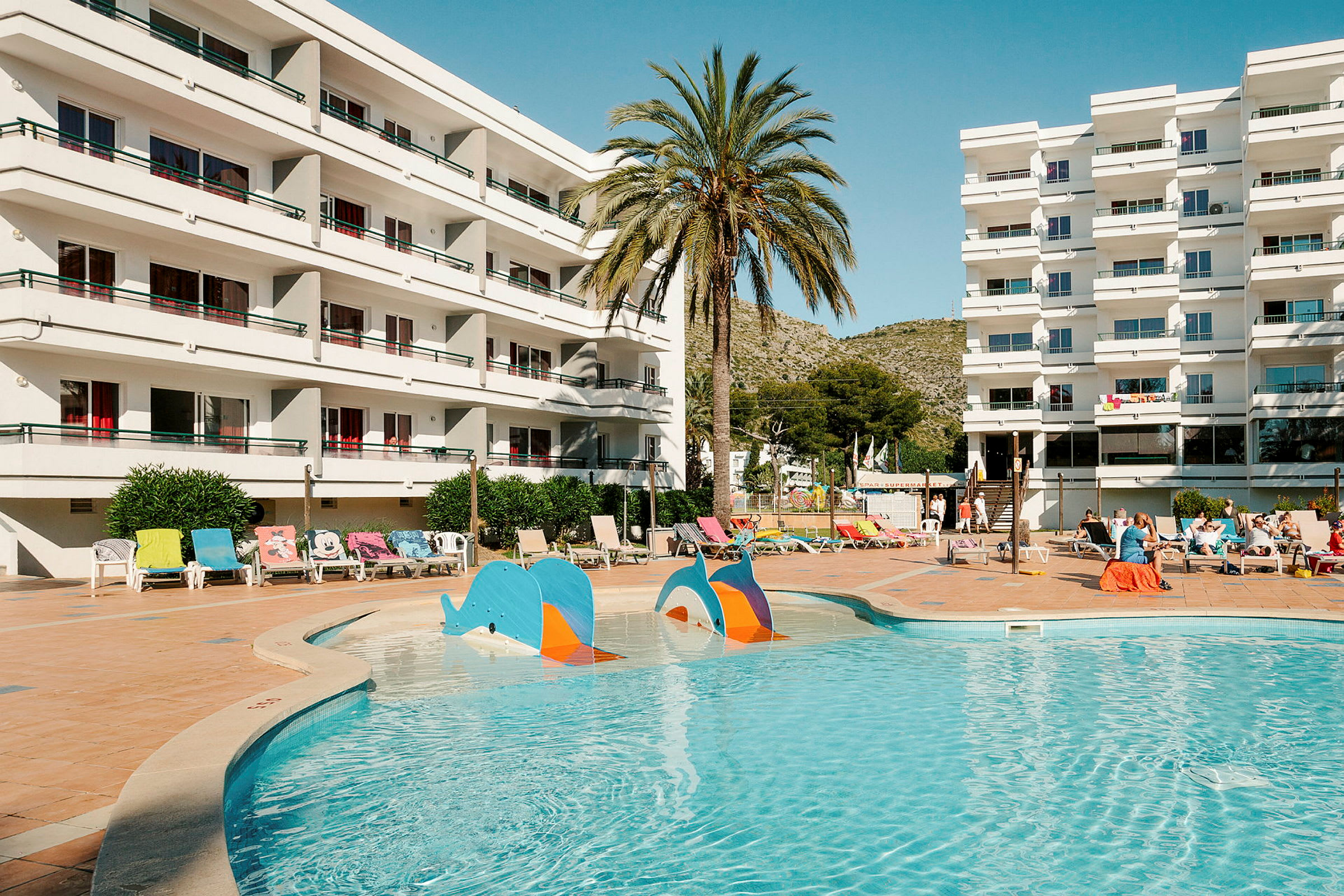 Hotellbyggnad med balkonger vid en utomhuspool och solstolar. (AI-genererad text)
