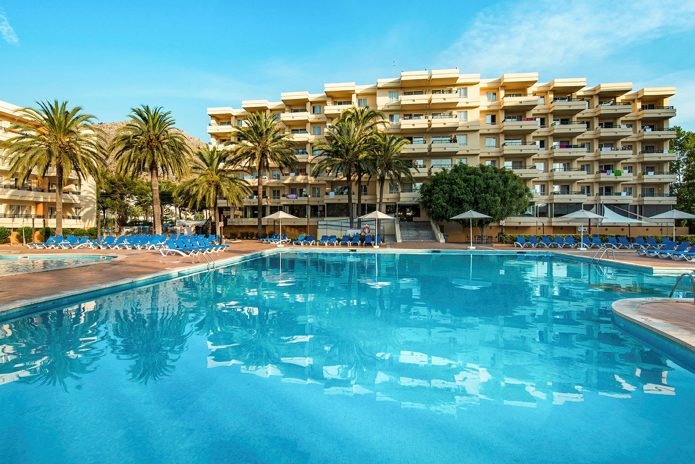En stor utomhuspool framför ett hotell med flera våningar omgiven av palmer. (AI-genererad text)