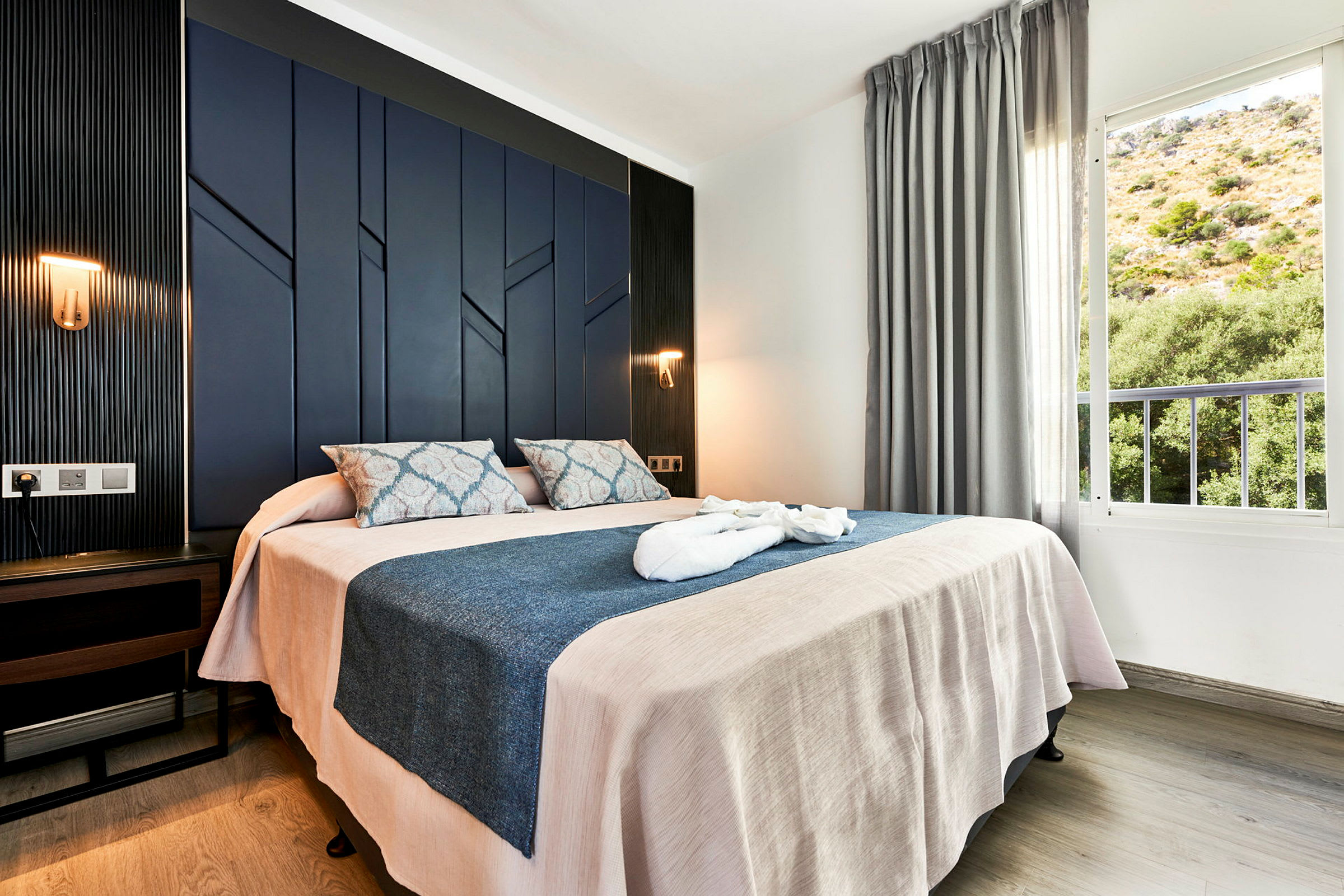 Hotellrum med dubbelbädd, mörkblå panelvägg och fönster med utsikt över grönskande område. (AI-genererad text)