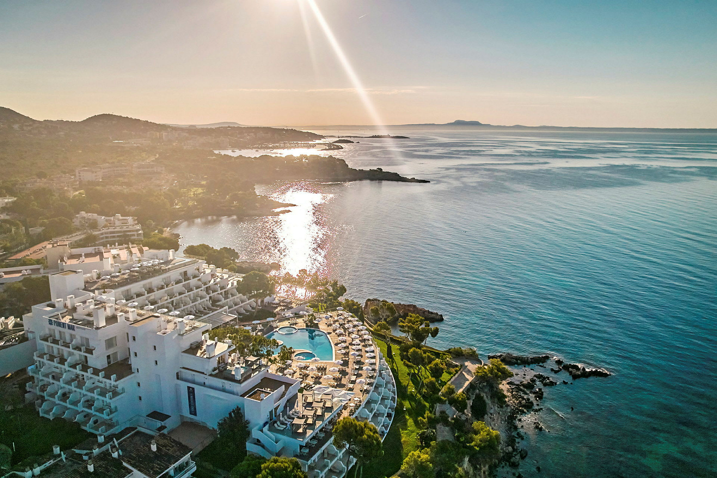 Flygvy av en kustnära hotellbyggnad med poolområde och utsikt över havet vid solnedgång. (AI-genererad text)