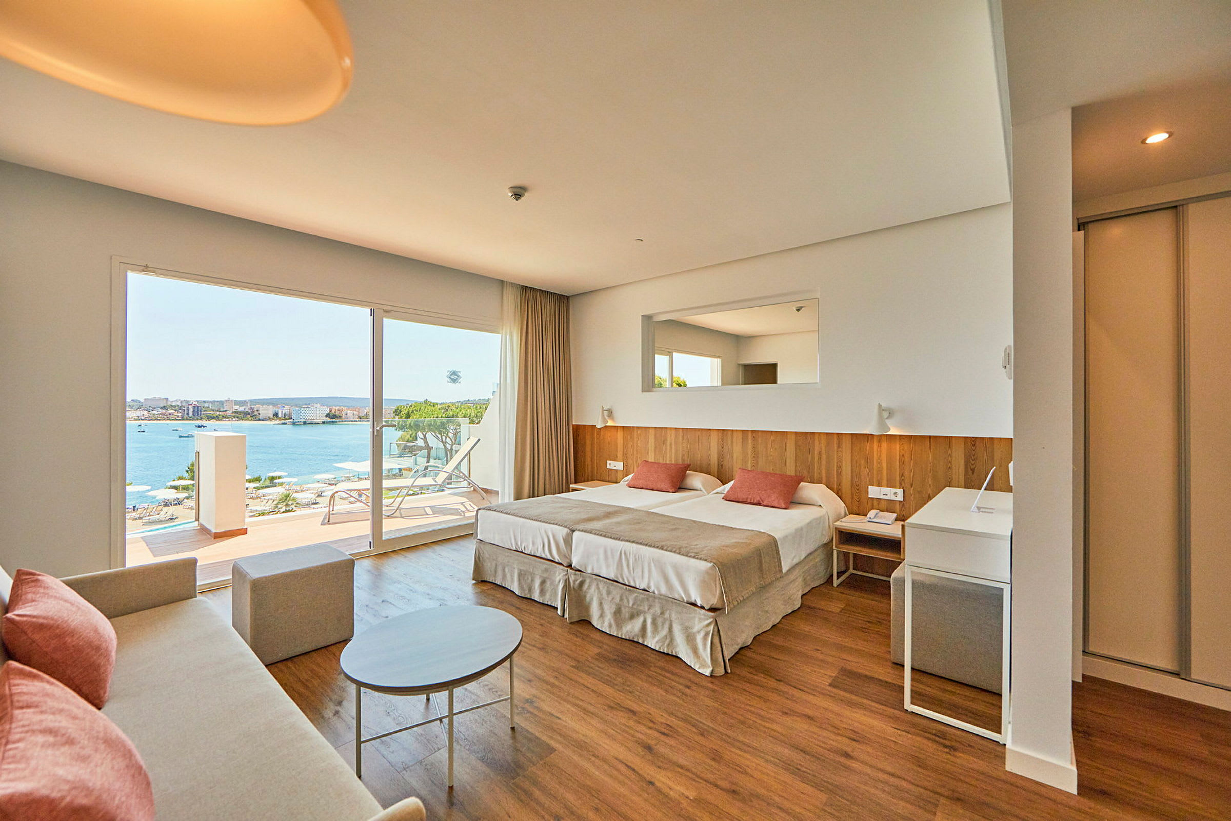 Ett hotellrum med dubbelsäng, soffa, och stora fönster som vetter mot en terrass med utsikt över havet. (AI-genererad text)