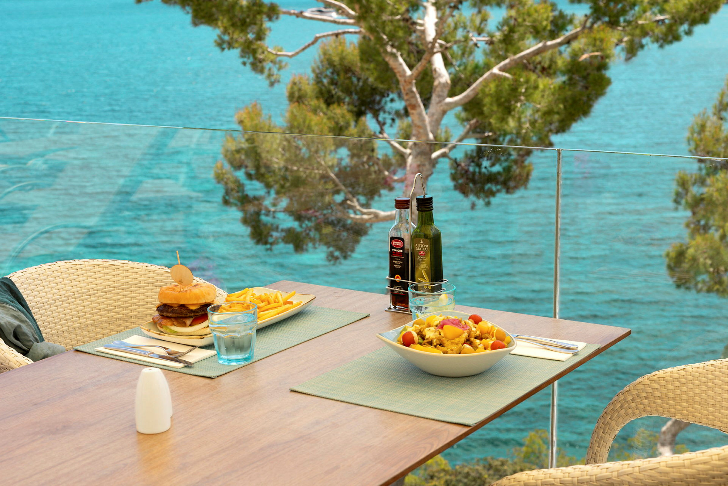 En terrass med utsikt över havet, där ett bord är uppdukat med en hamburgare, pommes frites och en sallad. (AI-genererad text)