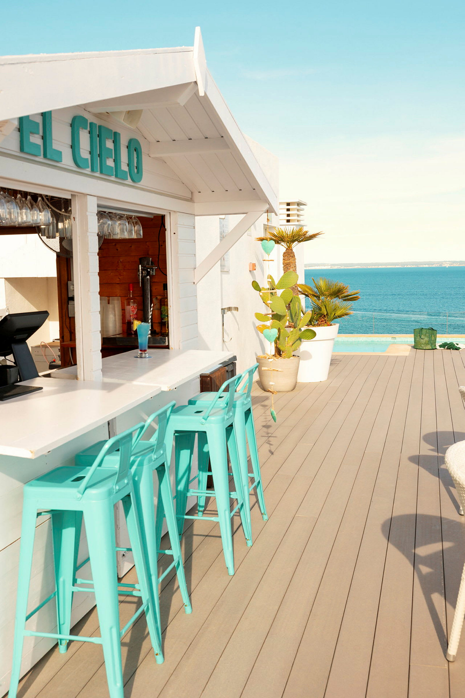 En strandbar med turkosa stolar vid en vit bar, med havet i bakgrunden. (AI-genererad text)