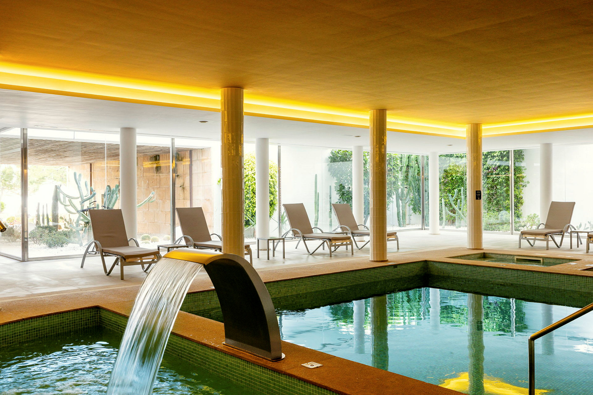 Inomhusområde med en uppvärmd pool och solstolar framför stora fönster ut mot en grönskande trädgård. (AI-genererad text)
