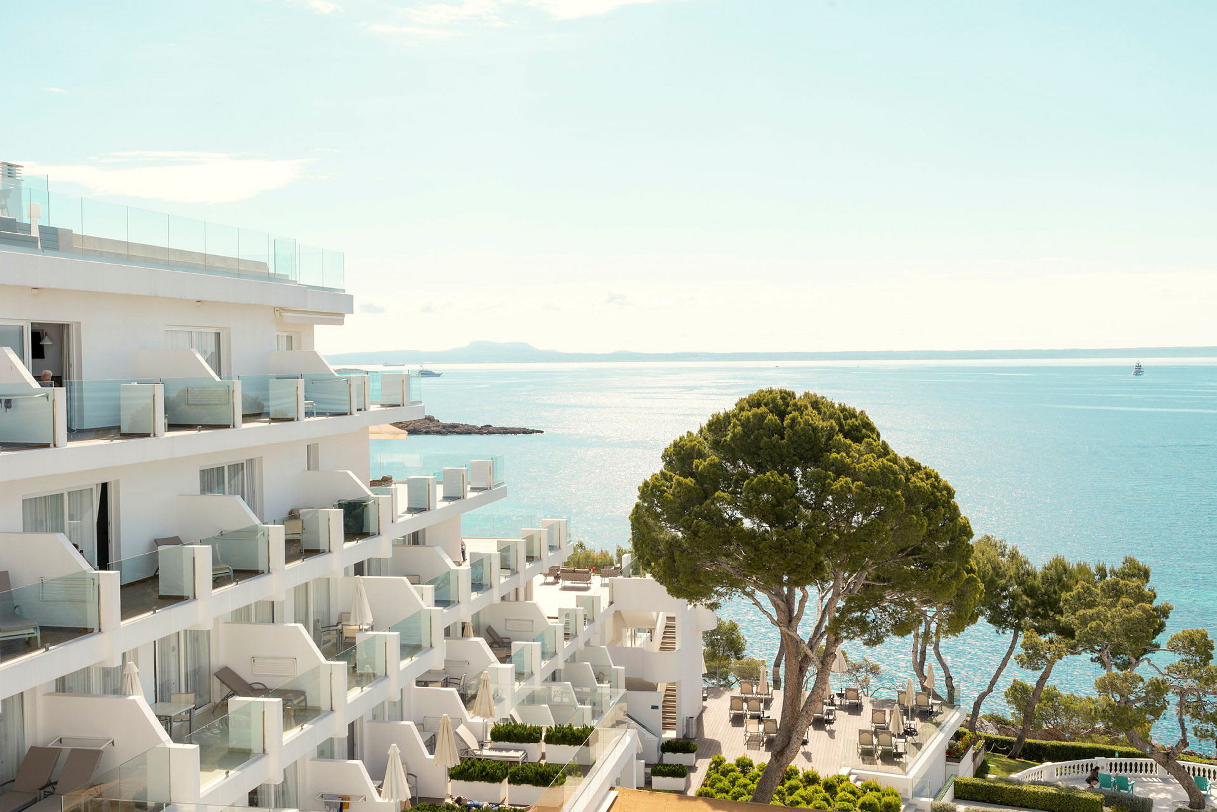Hotellbyggnad vid kusten med utsikt över havet och en terrass med bord och stolar samt träd. (AI-genererad text)