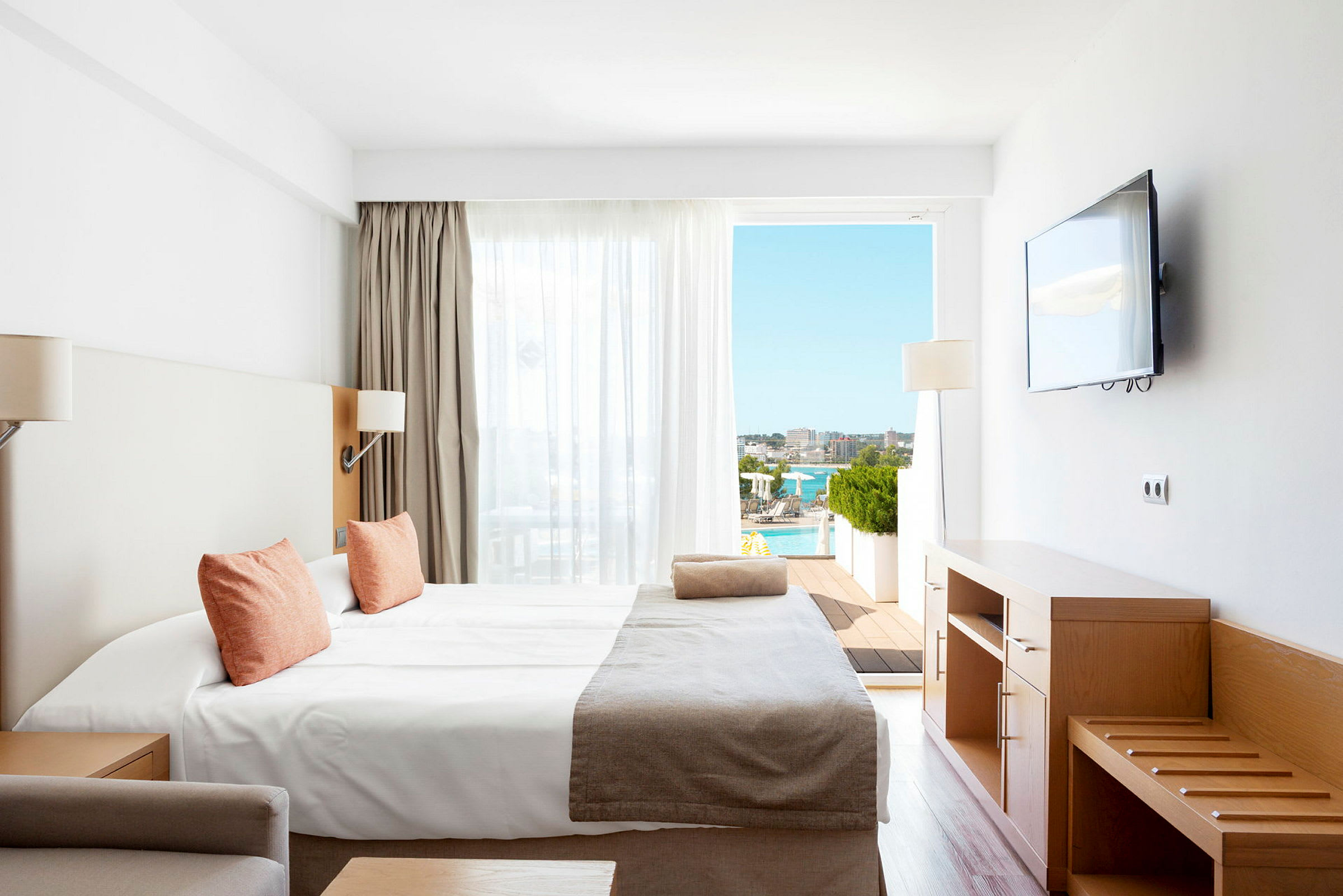 Ett hotellrum med en säng, träinredning, en väggmonterad TV och en utsikt över en solig terrass med pool. (AI-genererad text)