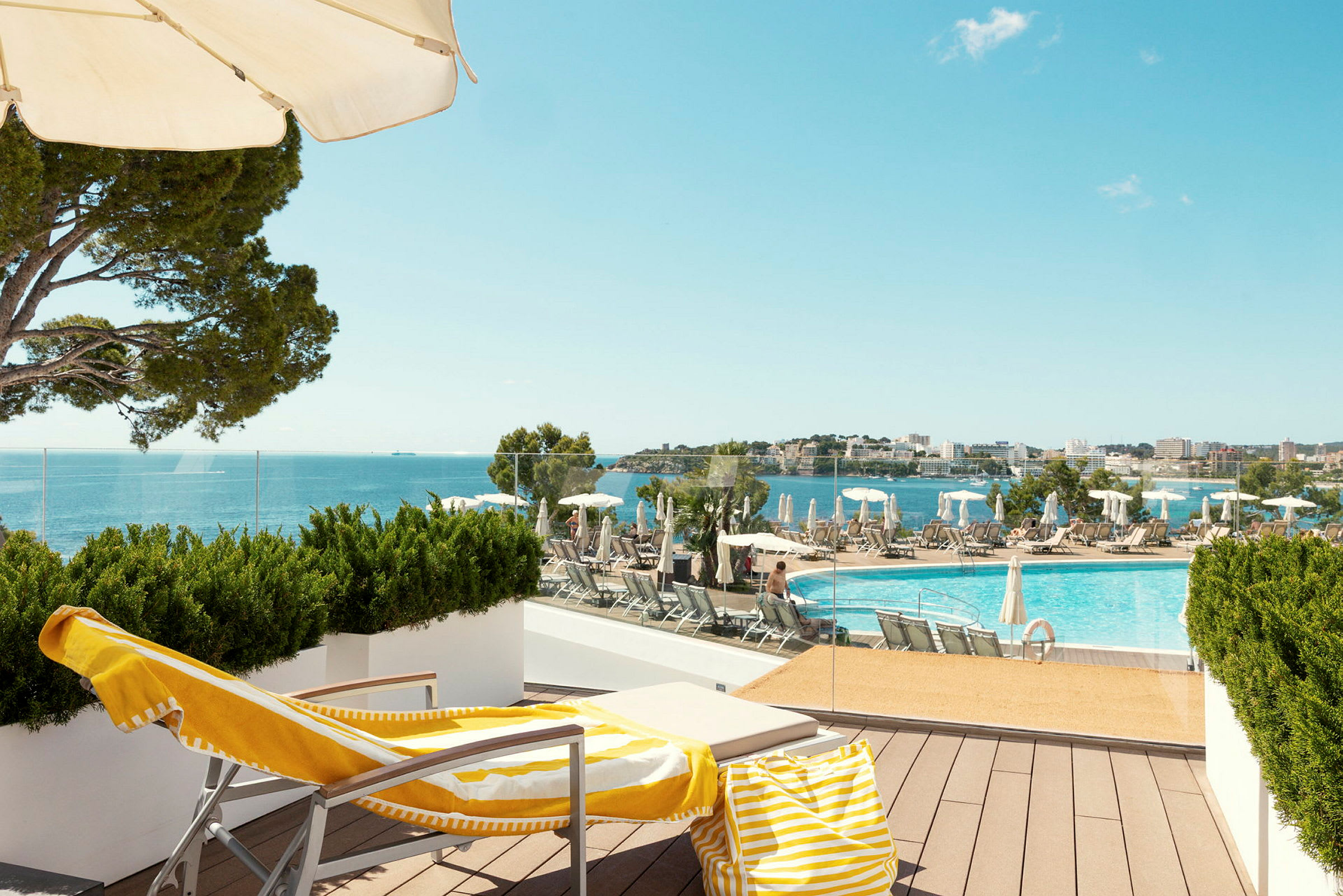 En solstol med gulvit handduk och strandväska står på en terrass med utsikt över en pool och havet. (AI-genererad text)