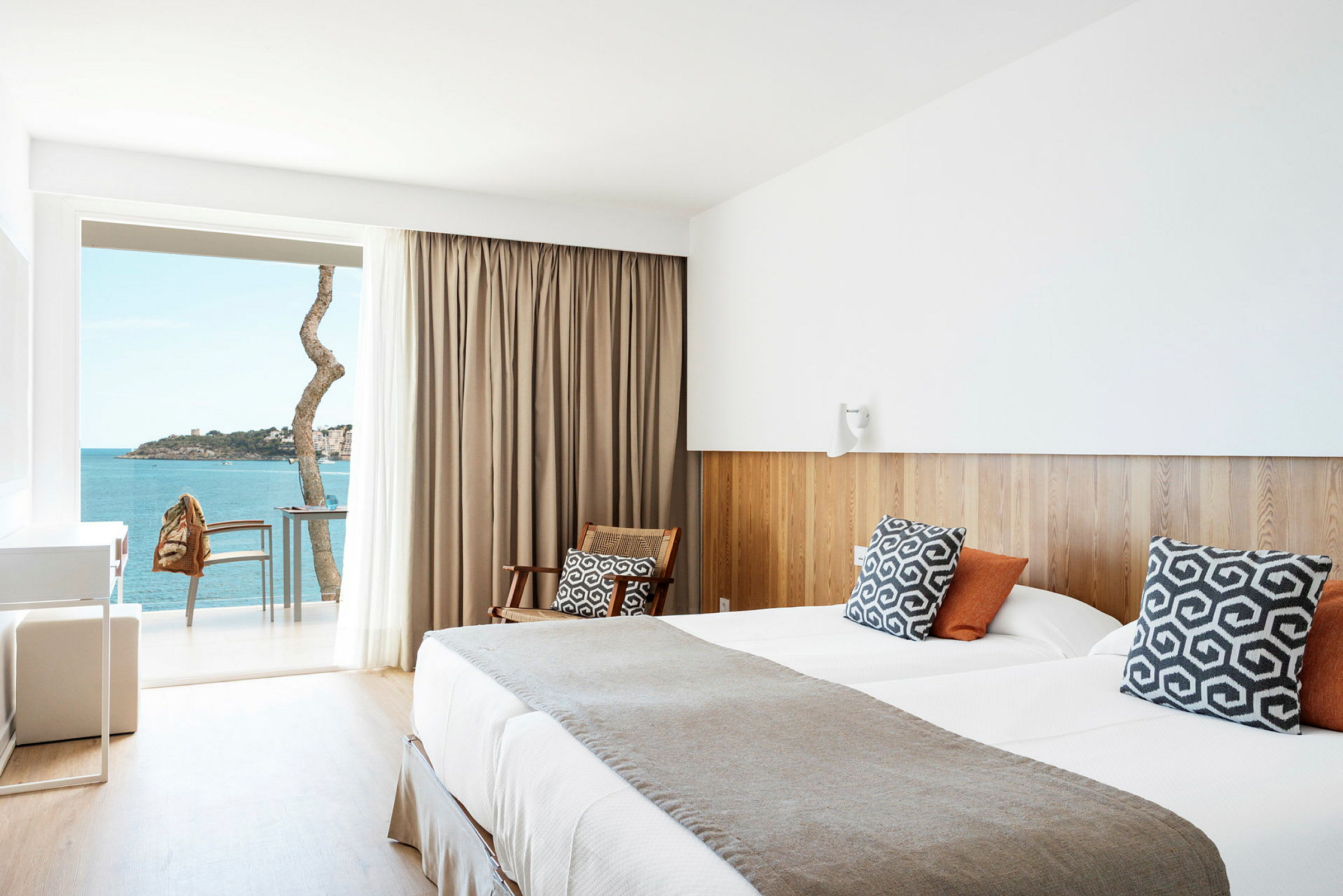 Ett hotellrum med moderna detaljer, en dubbelsäng, rustika kuddar, en sittplats och en balkong med utsikt över havet. (AI-genererad text)