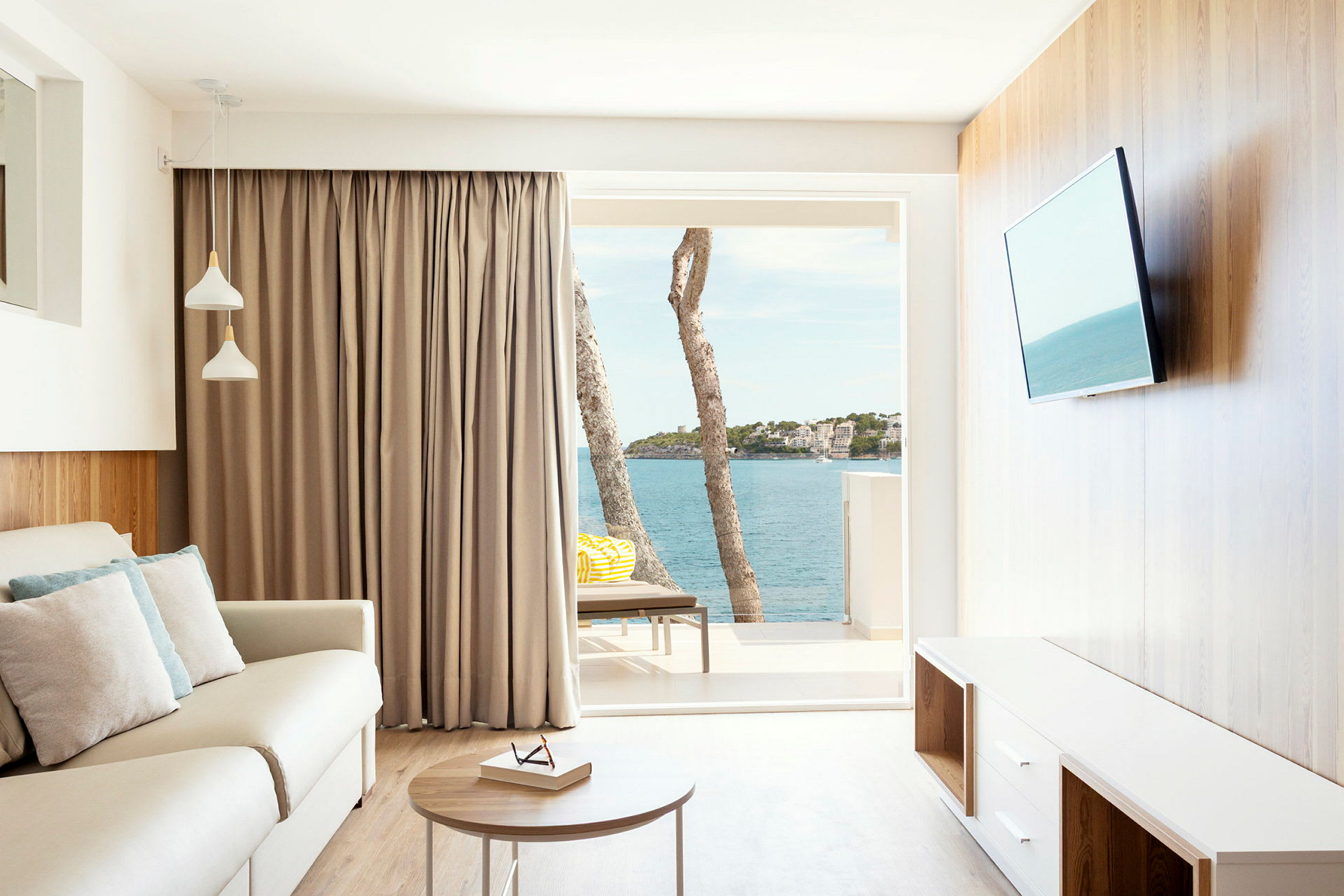 Ett ljust vardagsrum med en soffa, bord och TV. Fönstret visar en utsikt över havet och några träd. (AI-genererad text)