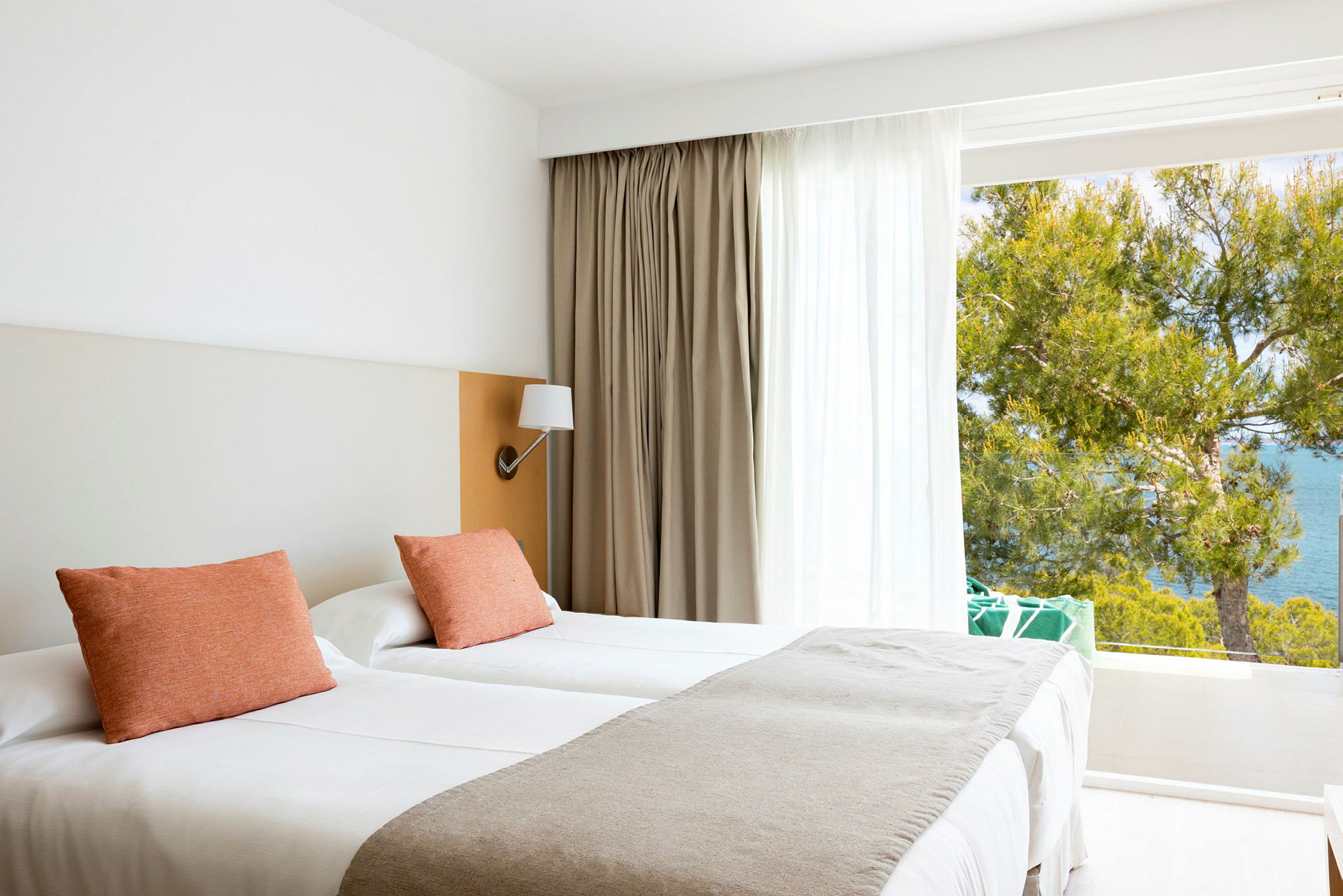 Ett ljust och modernt hotellrum med två enkelsängar, prydnadskuddar och utsikt mot havet genom stora fönster. (AI-genererad text)