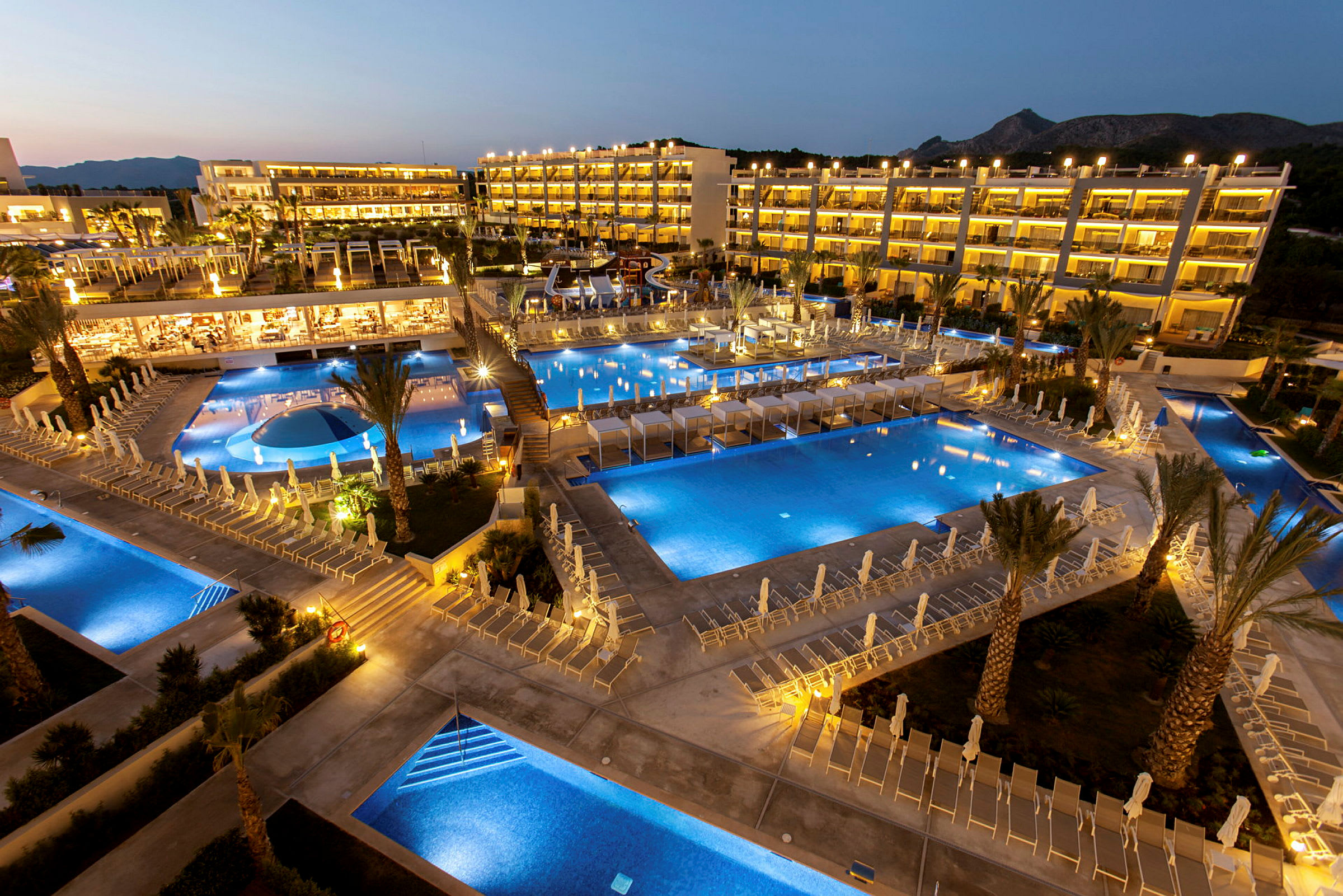 Ett hotellområde på kvällen med flera upplysta pooler och solstolar. (AI-genererad text)