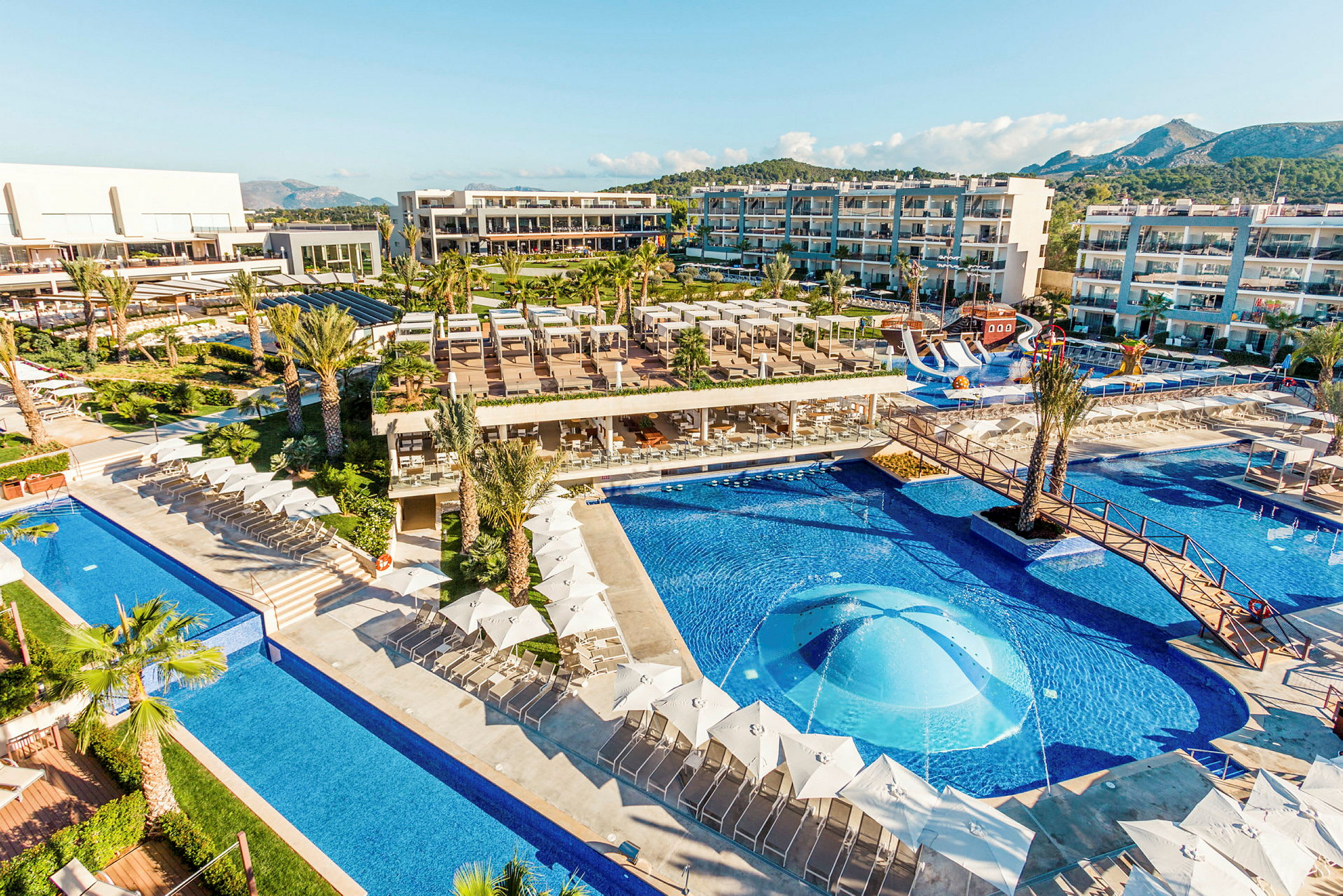 En vy över ett hotellområde med pooler, solstolar och byggnader. (AI-genererad text)