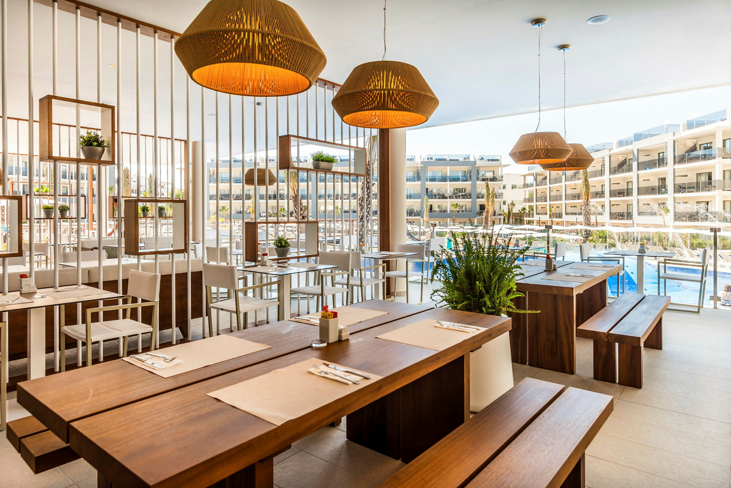 Restaurang med träbord och ljusa stolar, dekorerad med växter och lampor, med utsikt mot en pool och hotellbyggnader. (AI-genererad text)