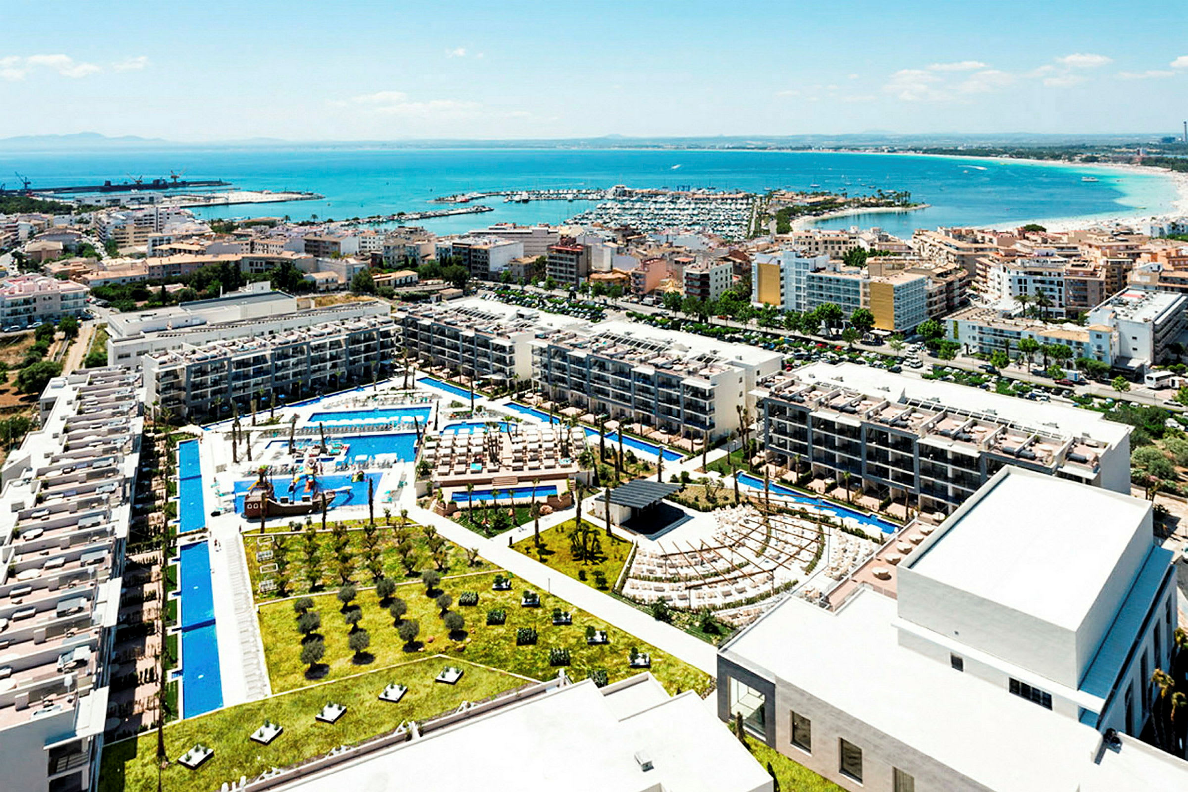 Utsikt över ett semesterresort vid kusten med pooler, byggnader och trädgårdar. Kustlinjen och havet syns i bakgrunden. (AI-genererad text)