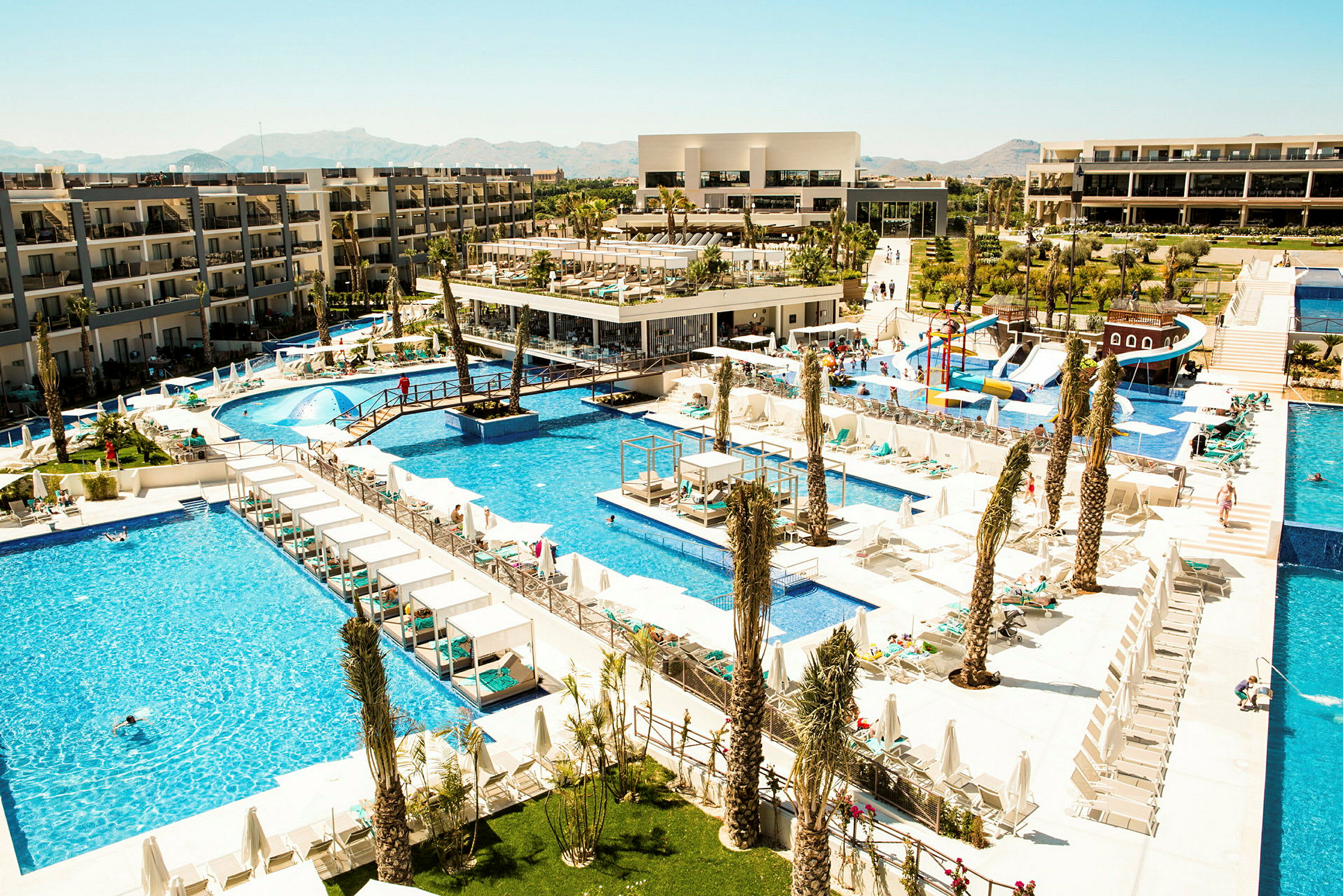 En stor hotellanläggning med flera poolområden. Solstolar och parasoller syns runt poolerna. (AI-genererad text)