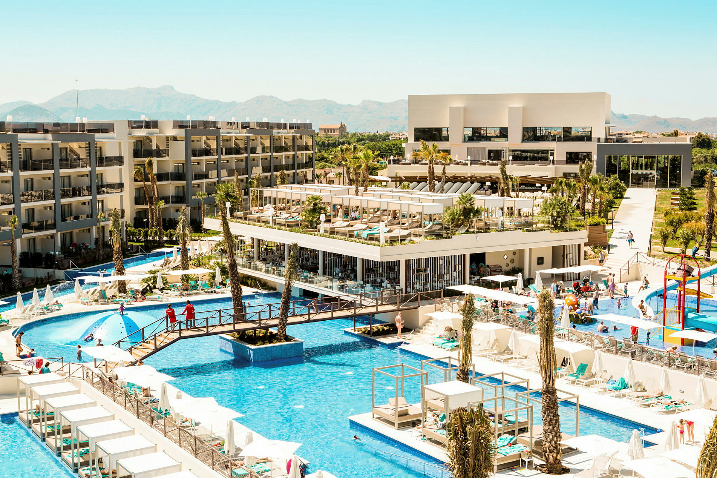 Vy över hotellområde med pooler, byggnader och solstolar. (AI-genererad text)