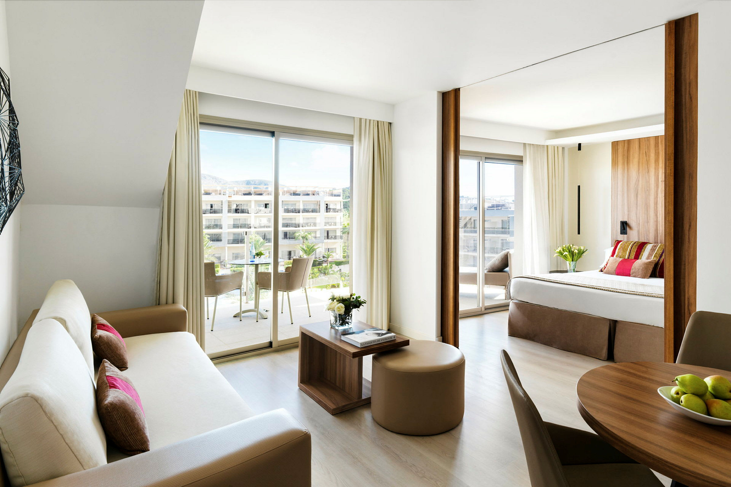 Ett modernt hotellrum med öppen planlösning, inkluderar ett vardagsrum med soffa och ett sovrum med dubbelsäng samt stora fönster med utsikt mot en terrass. (AI-genererad text)
