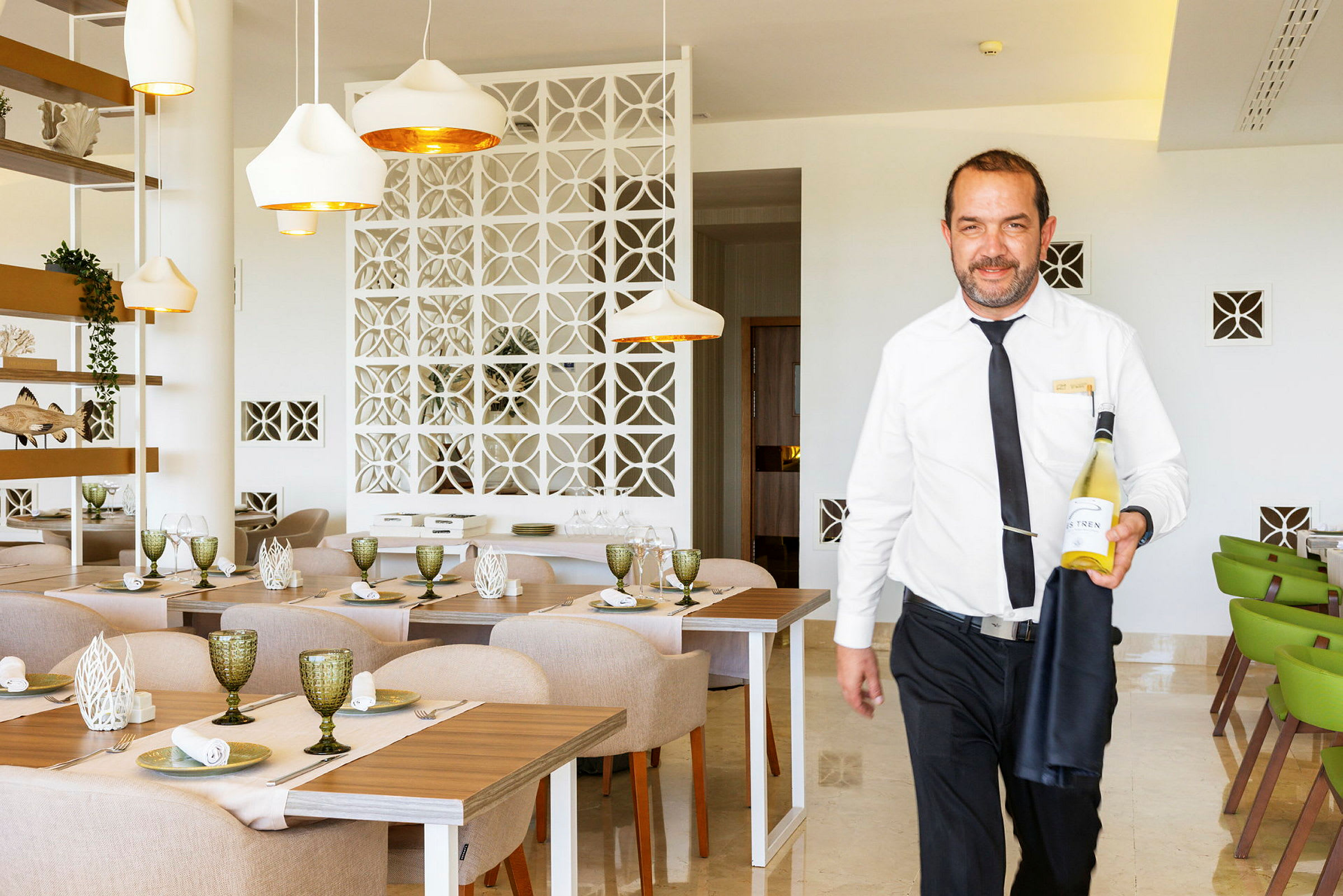 En servitör i vit skjorta håller en flaska vin i en ljus och modern restaurang. (AI-genererad text)