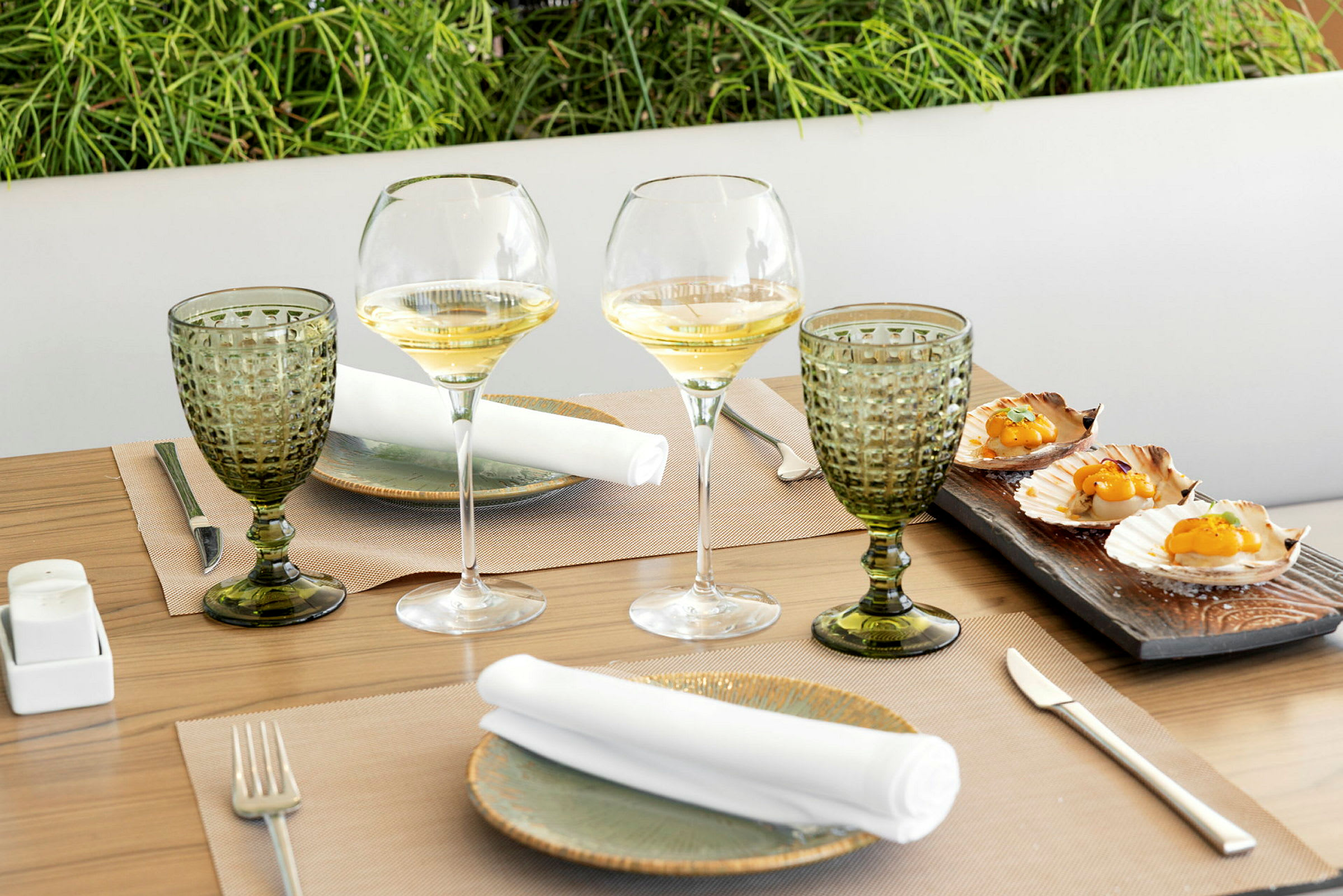 Ett dukat bord med vinglas, gröna dricksglas och tallrikar med vita servetter på. På en träplatta ligger tillagade pilgrimsmusslor. (AI-genererad text)