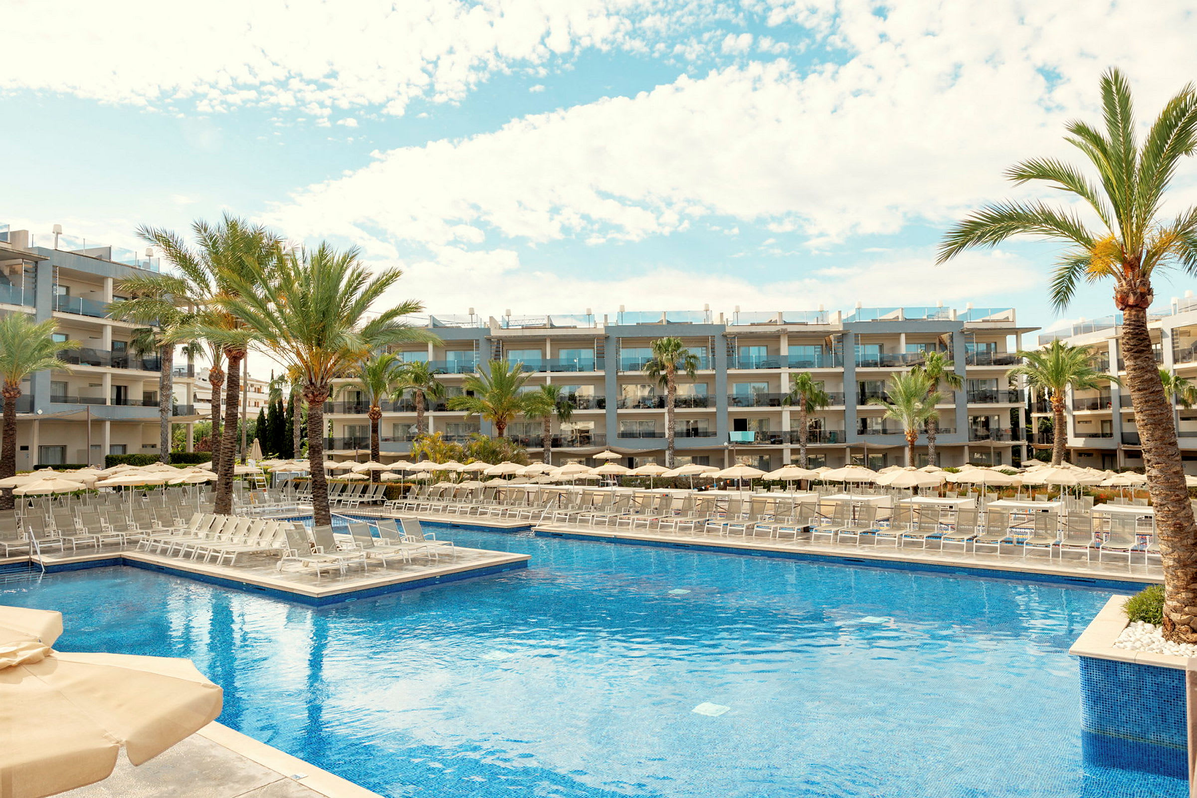 En bild på ett hotellområde med en stor pool omgiven av solstolar, parasoller och palmer. I bakgrunden syns moderna hotellbyggnader. (AI-genererad text)