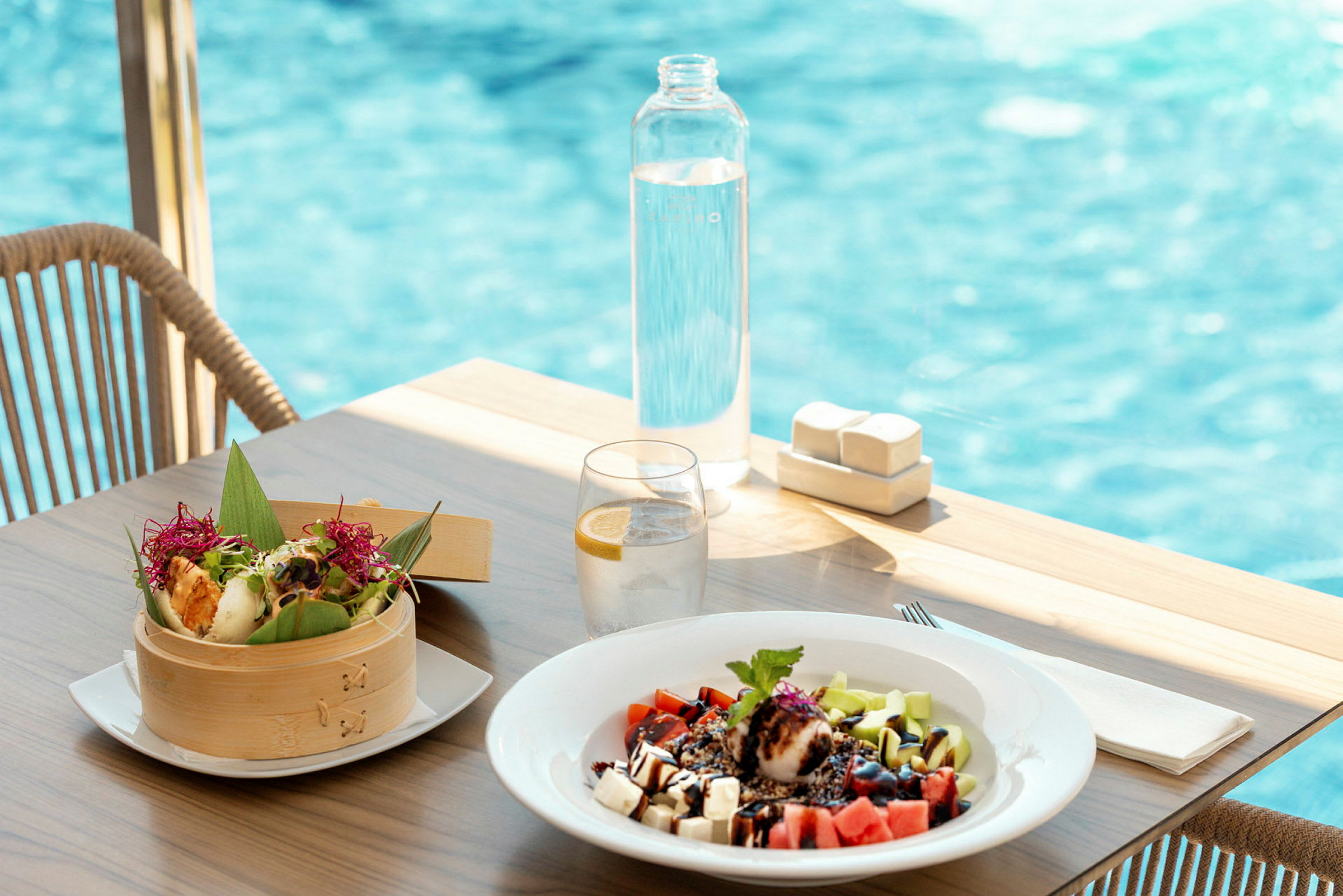 Ett dukat bord med en skål med färgglad mat, en tallrik med fruktsallad, ett glas vatten med citron och en flaska vatten. En pool syns i bakgrunden. (AI-genererad text)