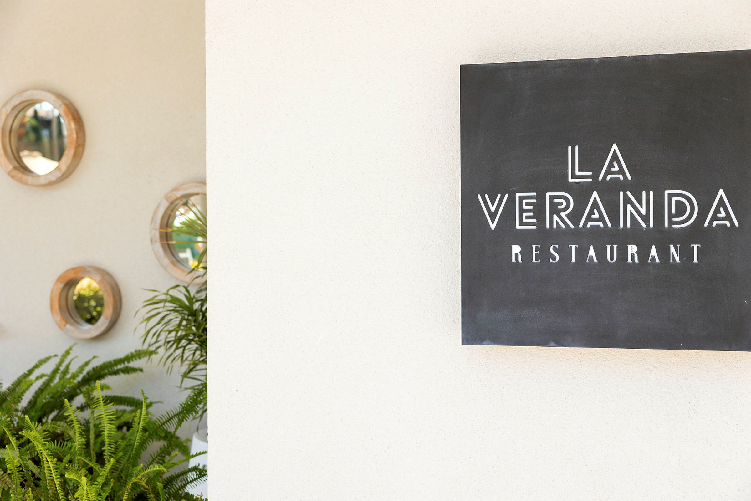 En skylt som visar texten "La Veranda Restaurant" bredvid en vägg dekorerad med runda speglar och gröna växter. (AI-genererad text)