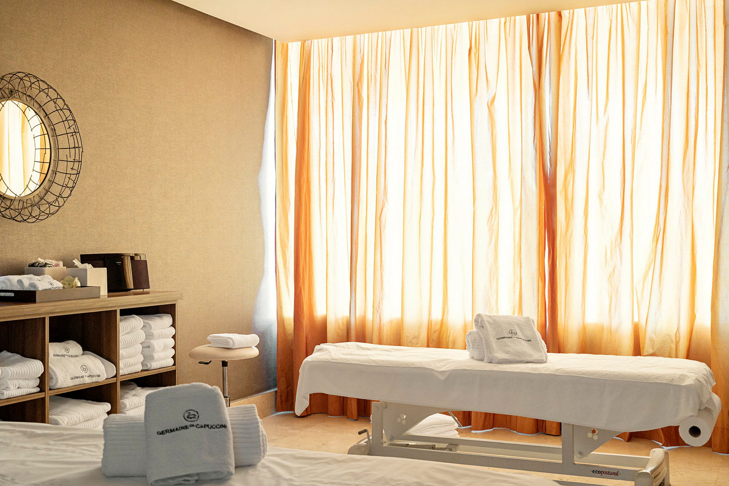 Ett ljust rum med massagebänkar och handdukar, dekorerat med ett runt väggspegel och skåp fyllda med handdukar. (AI-genererad text)