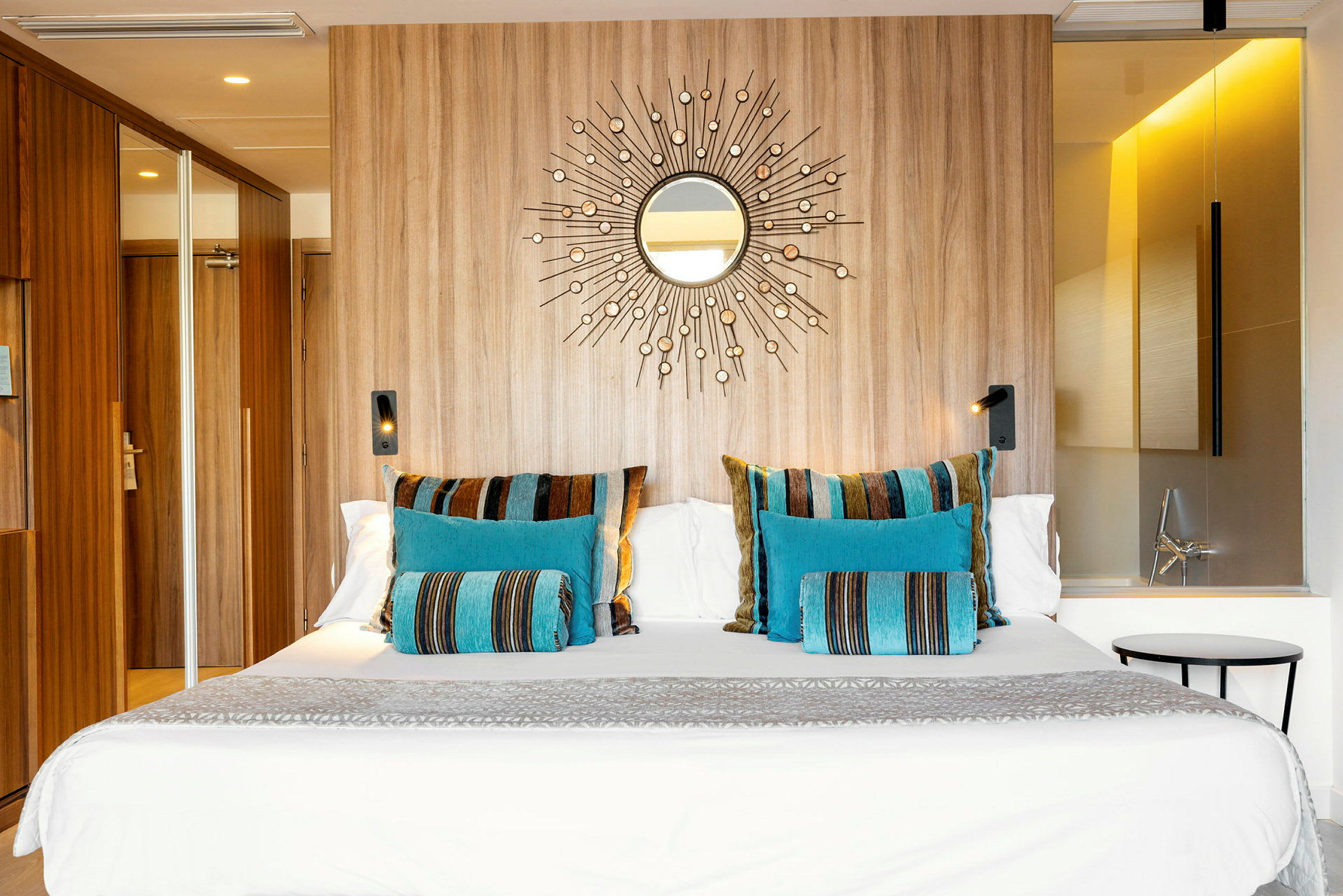 Ett hotellrum med en stor säng dekorerad med färgglada kuddar i blått och randigt mönster, en trävägg med ett spegelmotiv och modern belysning. (AI-genererad text)