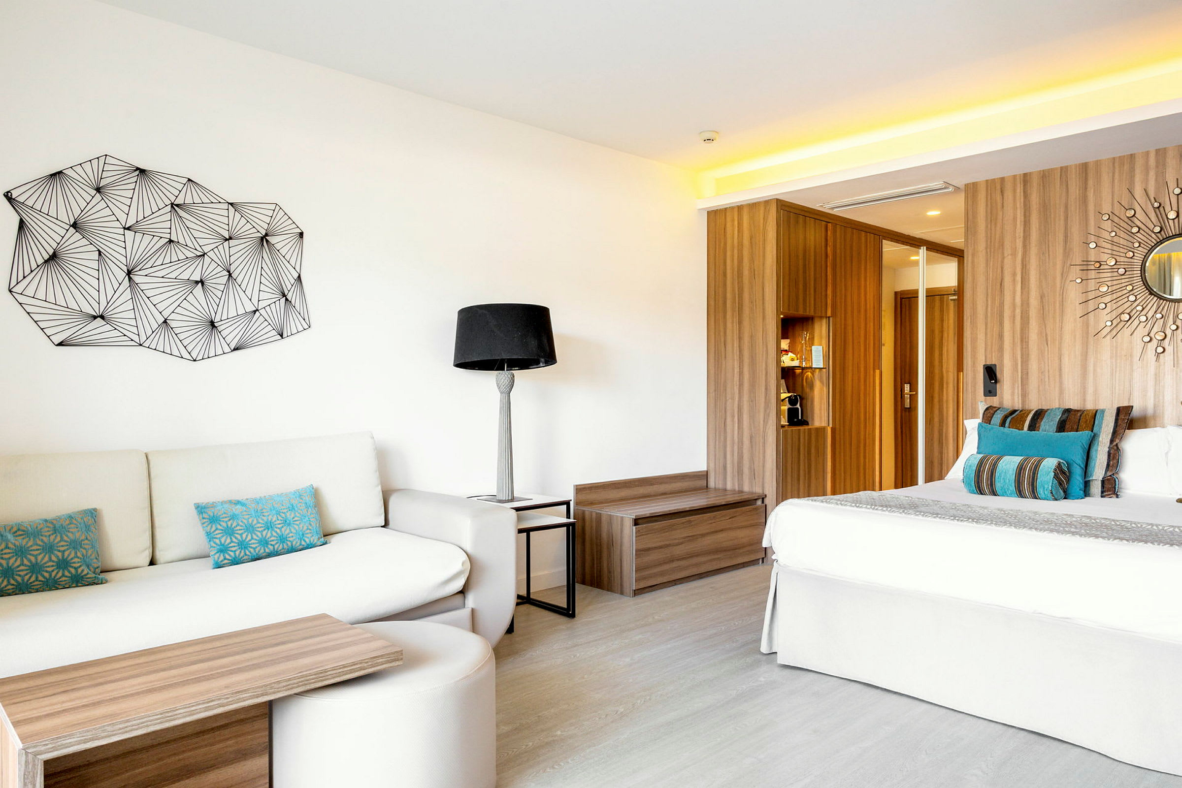 Ett modernt hotellrum med en soffa, färgglada kuddar, ett soffbord, en säng med dekorativa kuddar samt en träpanel bakom. På väggen finns konst och ett litet runt spegeldekoration. (AI-genererad text)