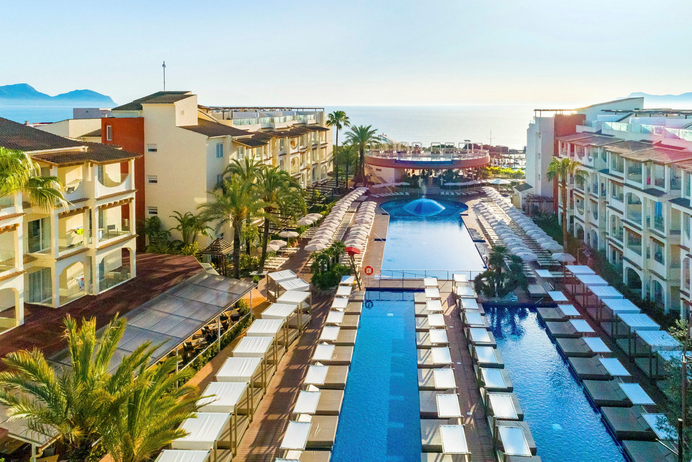 Ett hotellområde med en stor pool, solstolar och parasoller, omgiven av byggnader med balkonger. (AI-genererad text)