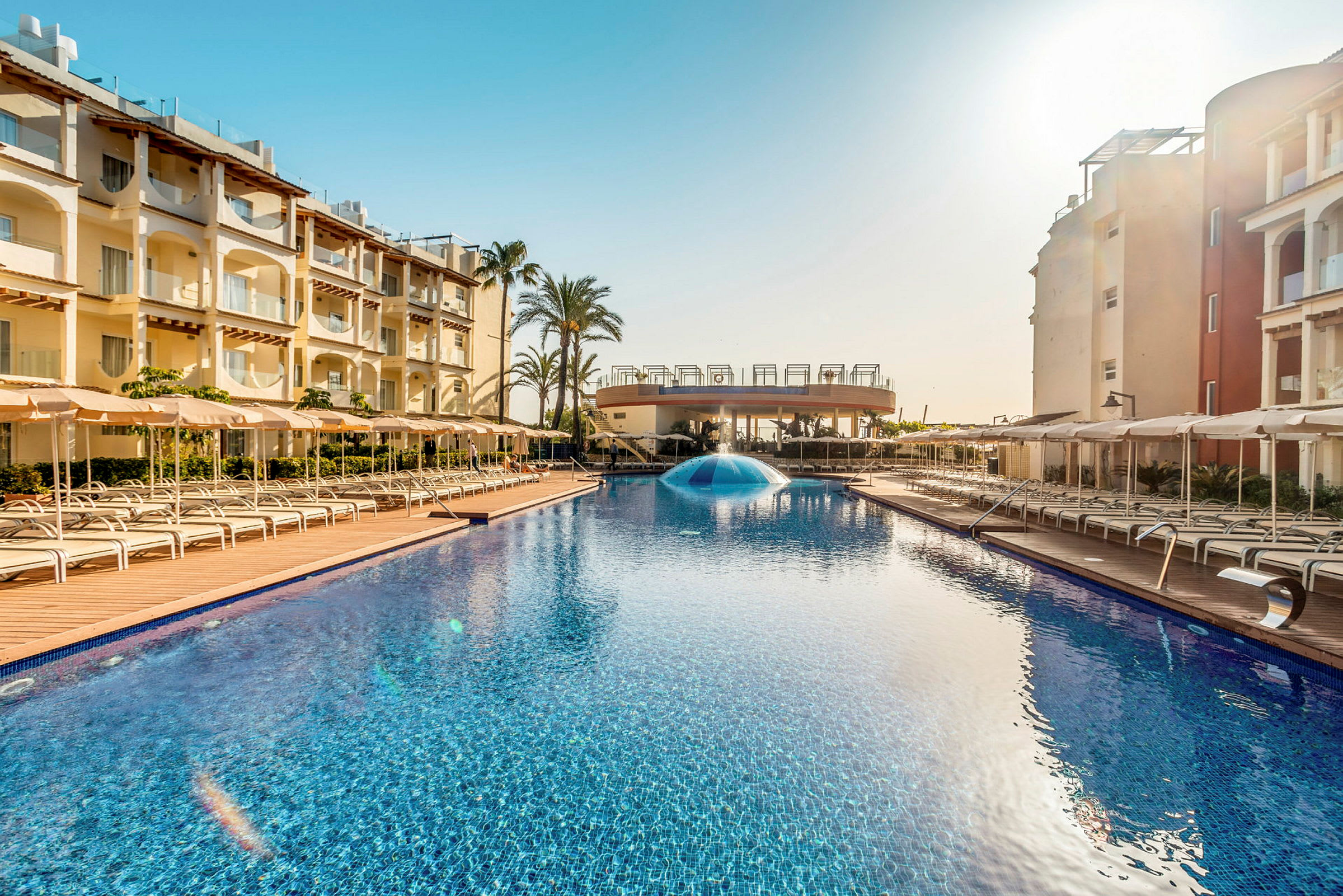 Utomhuspool vid hotell omgivet av solstolar och byggnader. (AI-genererad text)