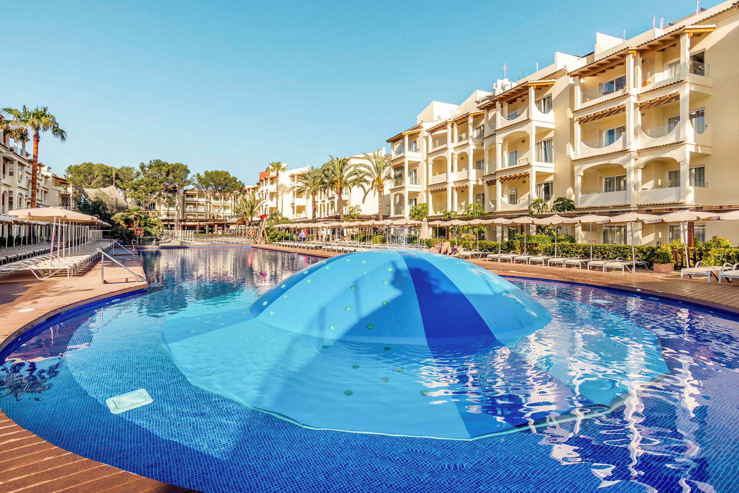 Poolområde med blått vatten och hotellbyggnad i bakgrunden. (AI-genererad text)