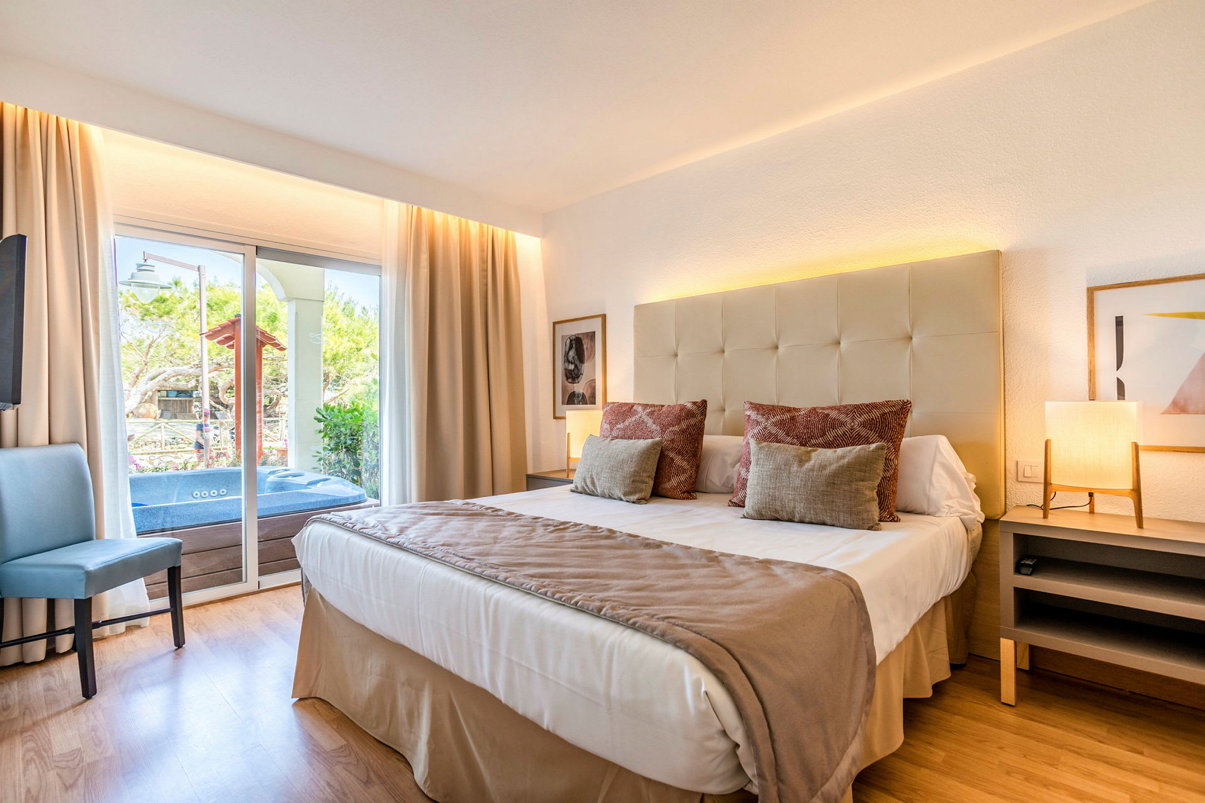 Modernt hotellrum med säng, lampor och utsikt till en terrass med grönska och jacuzzi. (AI-genererad text)